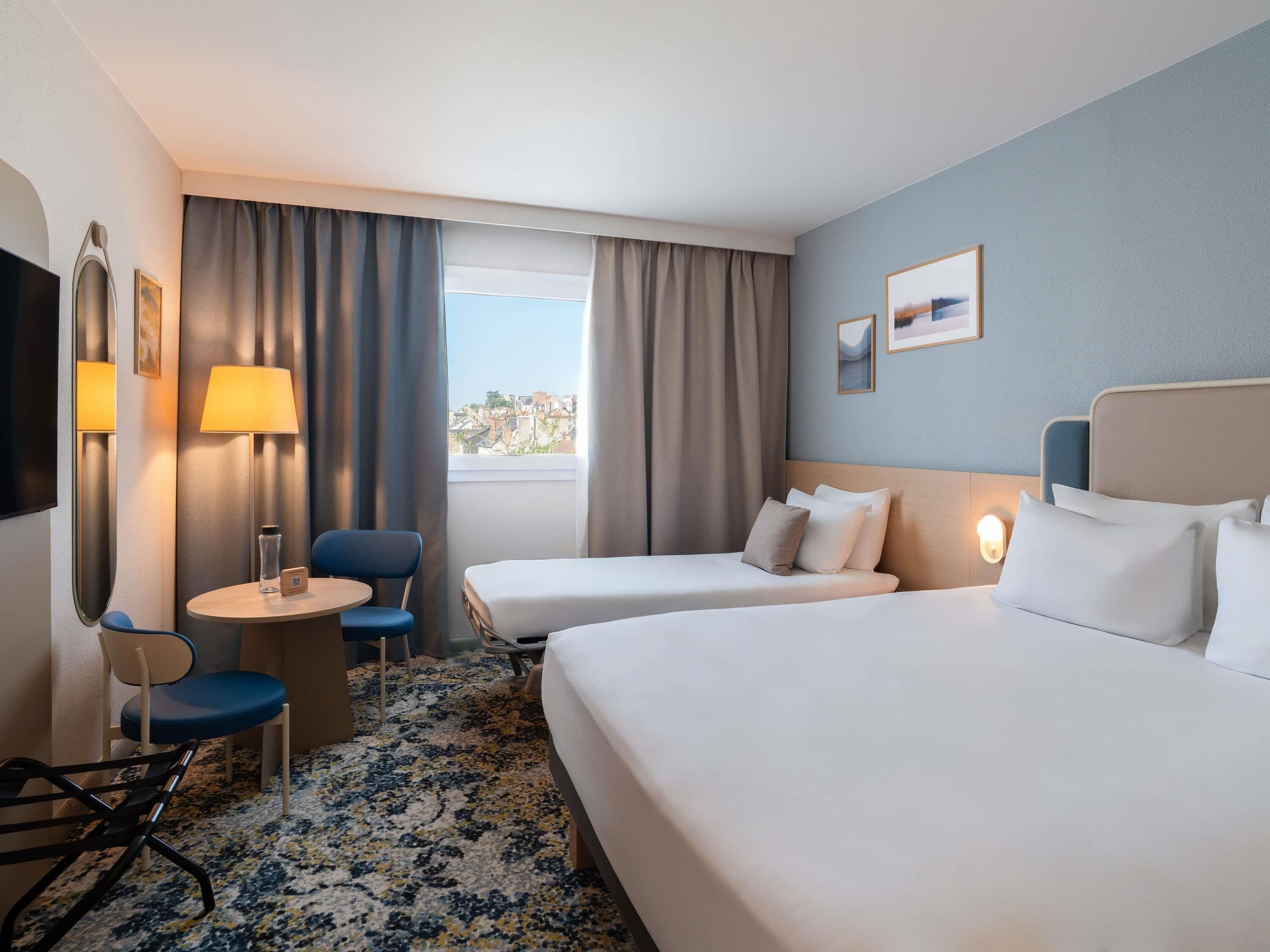 Habitación de hotel con dos camas, un escritorio, una silla, una lámpara y vistas a edificios a través del ventanal.