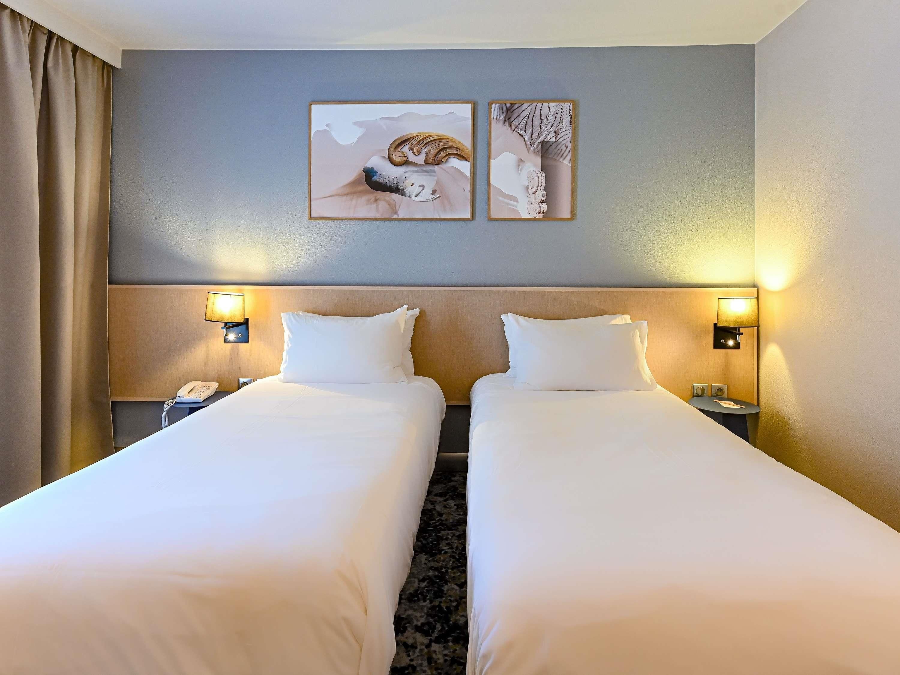 A hotel room with two beds, a headboard, bedside lamps, a phone, and two framed pictures on the wall.