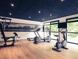 Une salle de sport équipée de machines de cardio, d’appareils de musculation et de grandes fenêtres qui laissent entrer la lumière naturelle et offrent une vue sur les arbres.