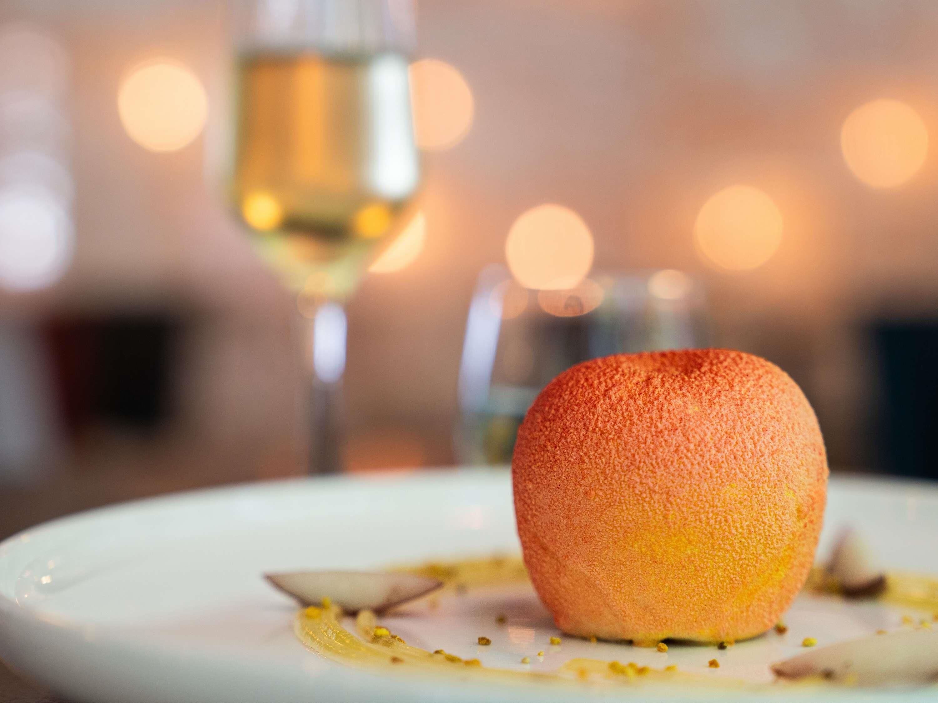 Ein Dessert mit einer kugelförmigen, orangen Komponente auf einem Teller, serviert mit einem Glas Weißwein.