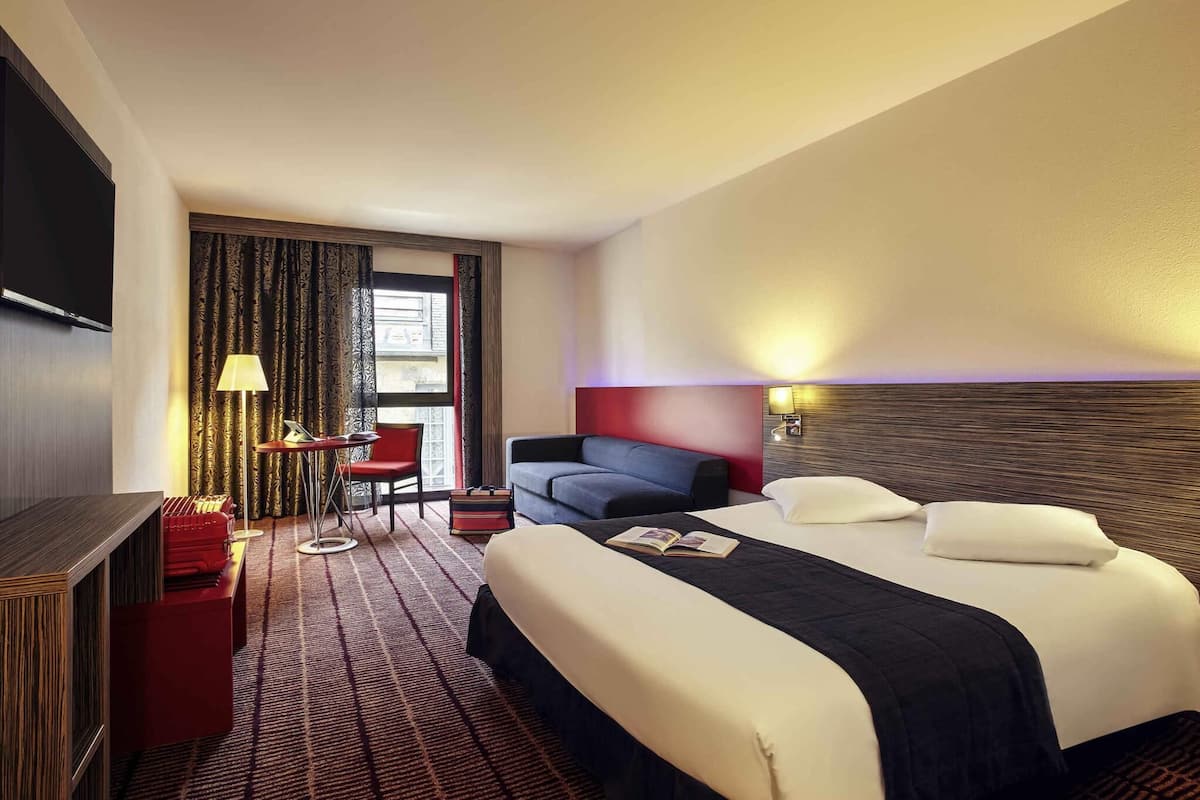 A hotel room with a bed, a desk with a chair, a TV, and a window with curtains.