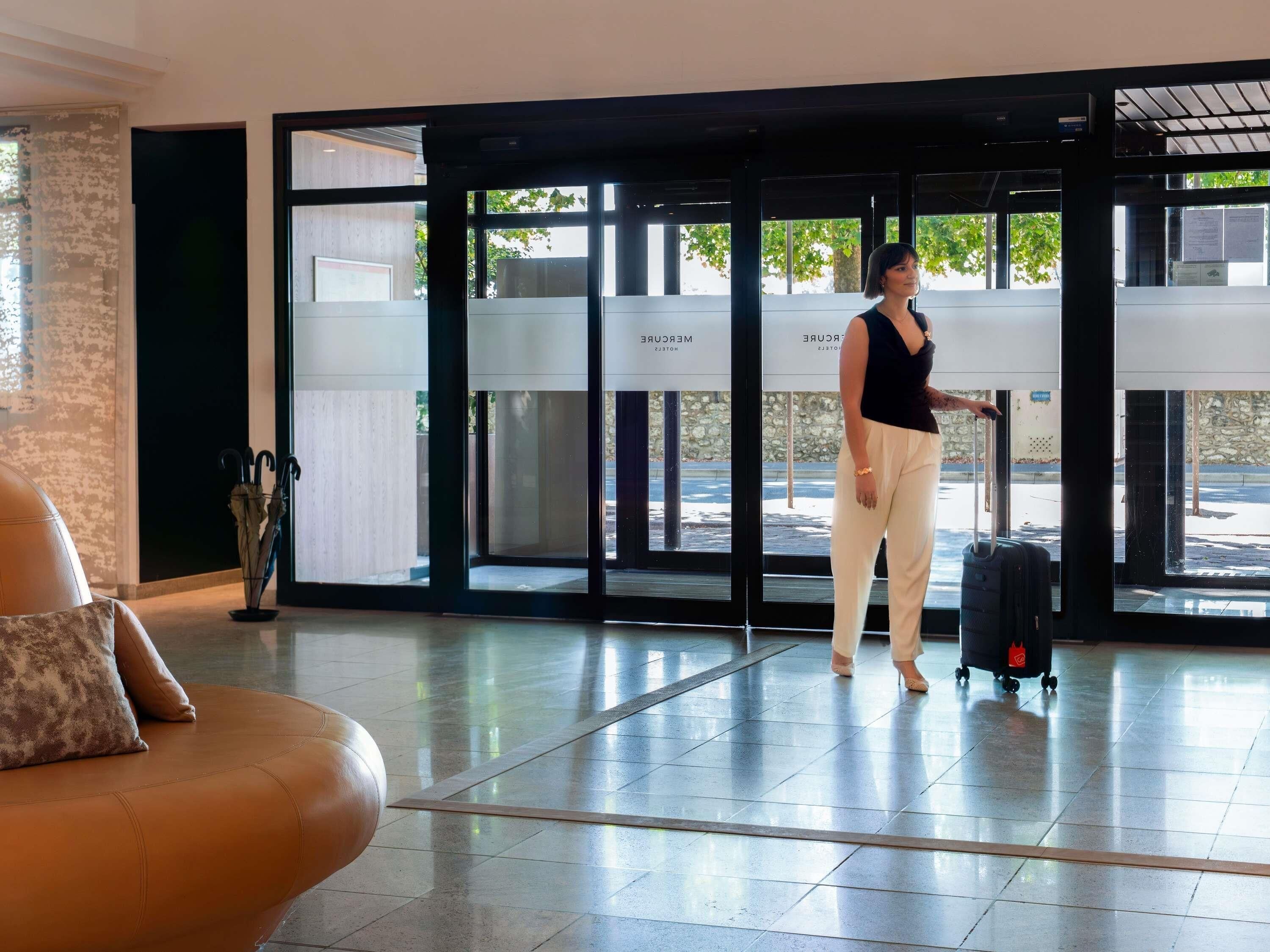 A person standing in a lobby with a suitcase, large glass doors, and a seating area with a cushion.