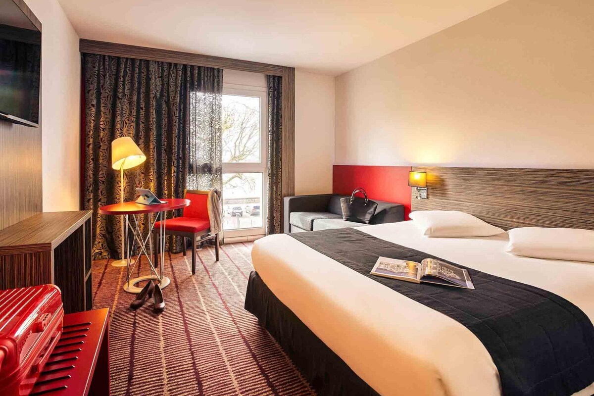 Quarto de hotel com cama, escrivaninha com abajur, cadeira, sofá e televisão.