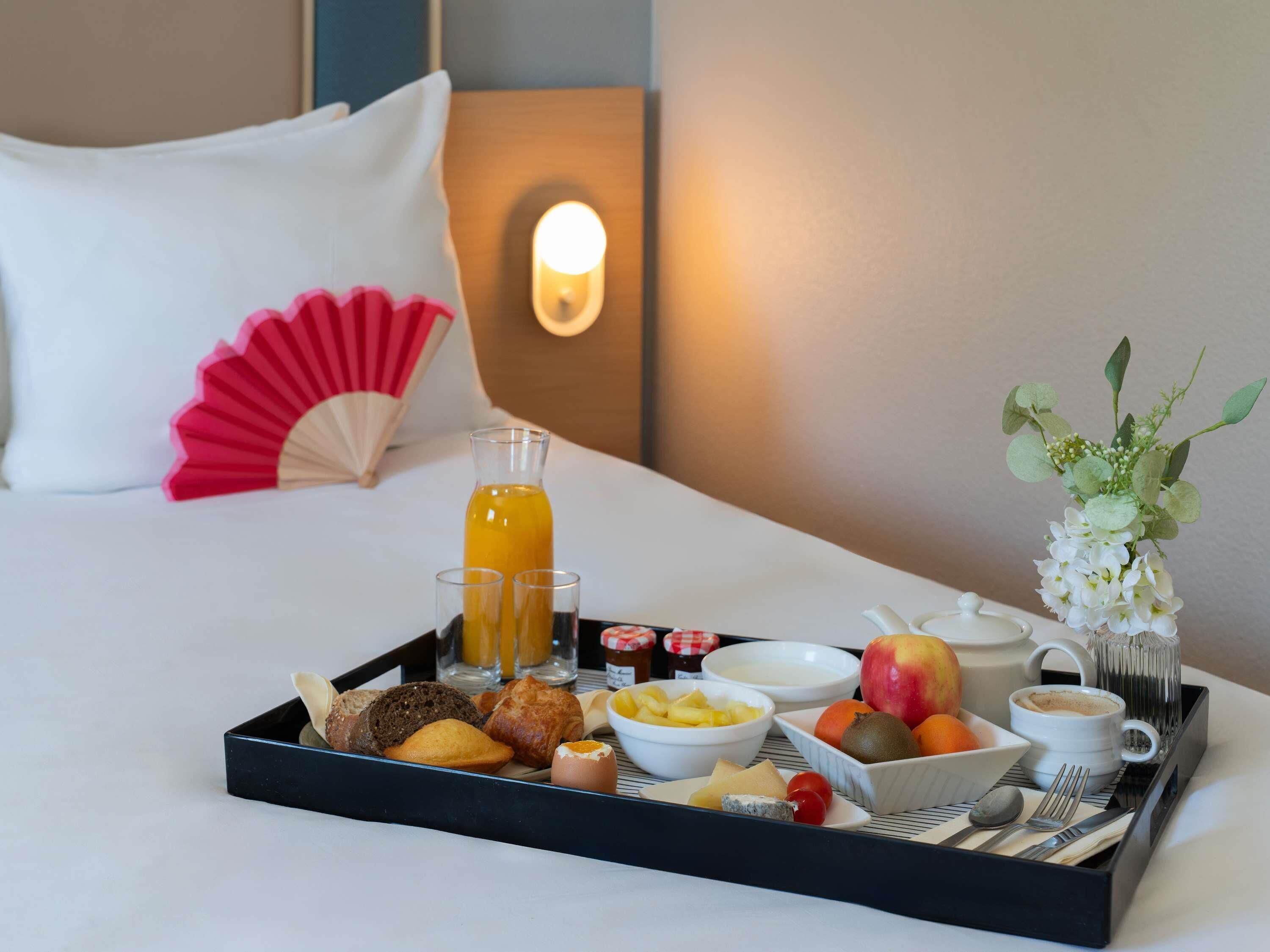 A neatly arranged breakfast tray on a bed with a pillow, a bedside lamp, and a vase of flowers.