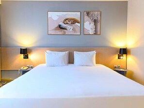 A hotel room with a large bed, two bedside lamps, a telephone, and two framed artworks on the wall.