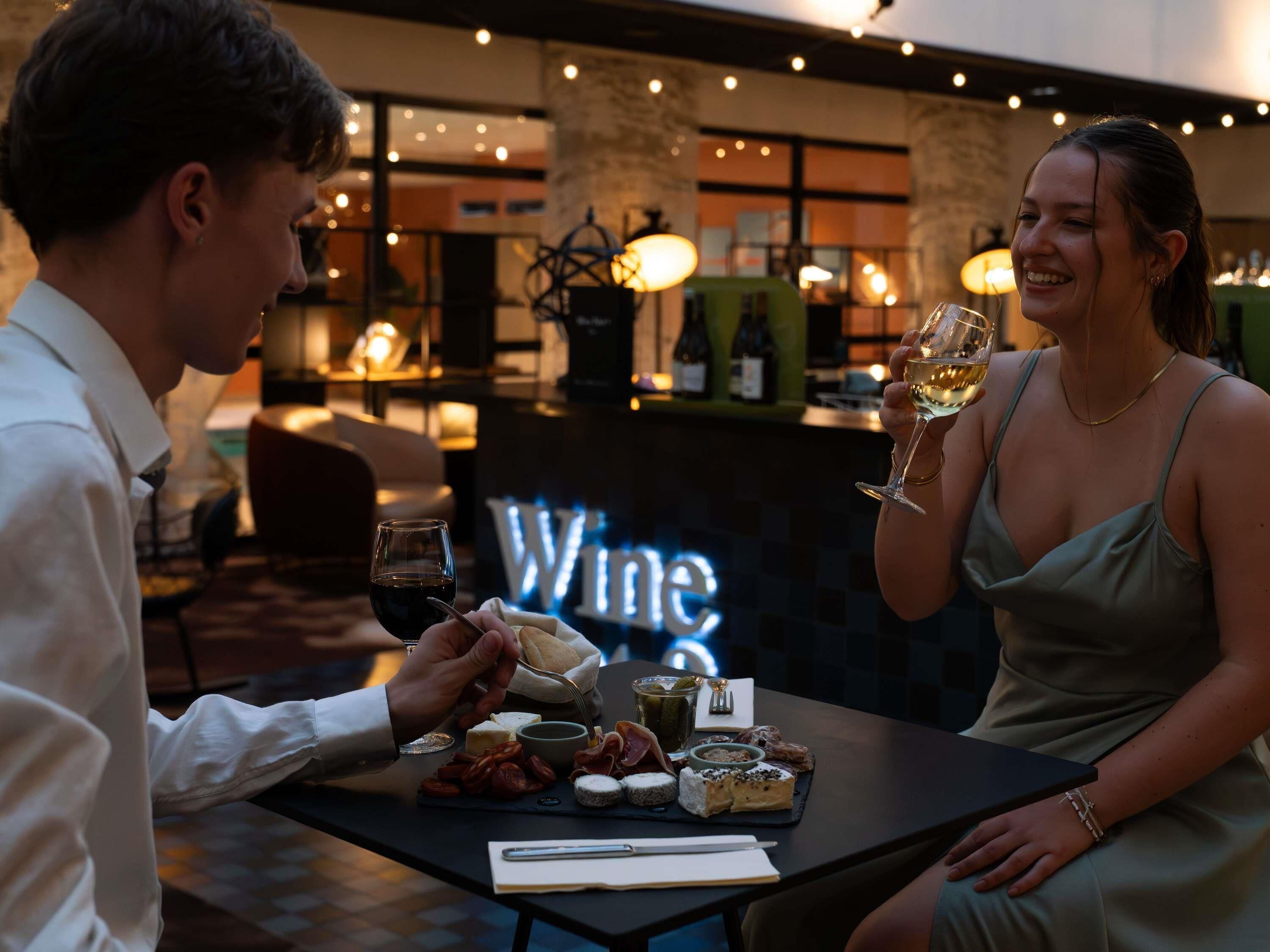 Una pareja sentada en una mesa con una copa de vino, una botella y una tabla de quesos y embutidos.