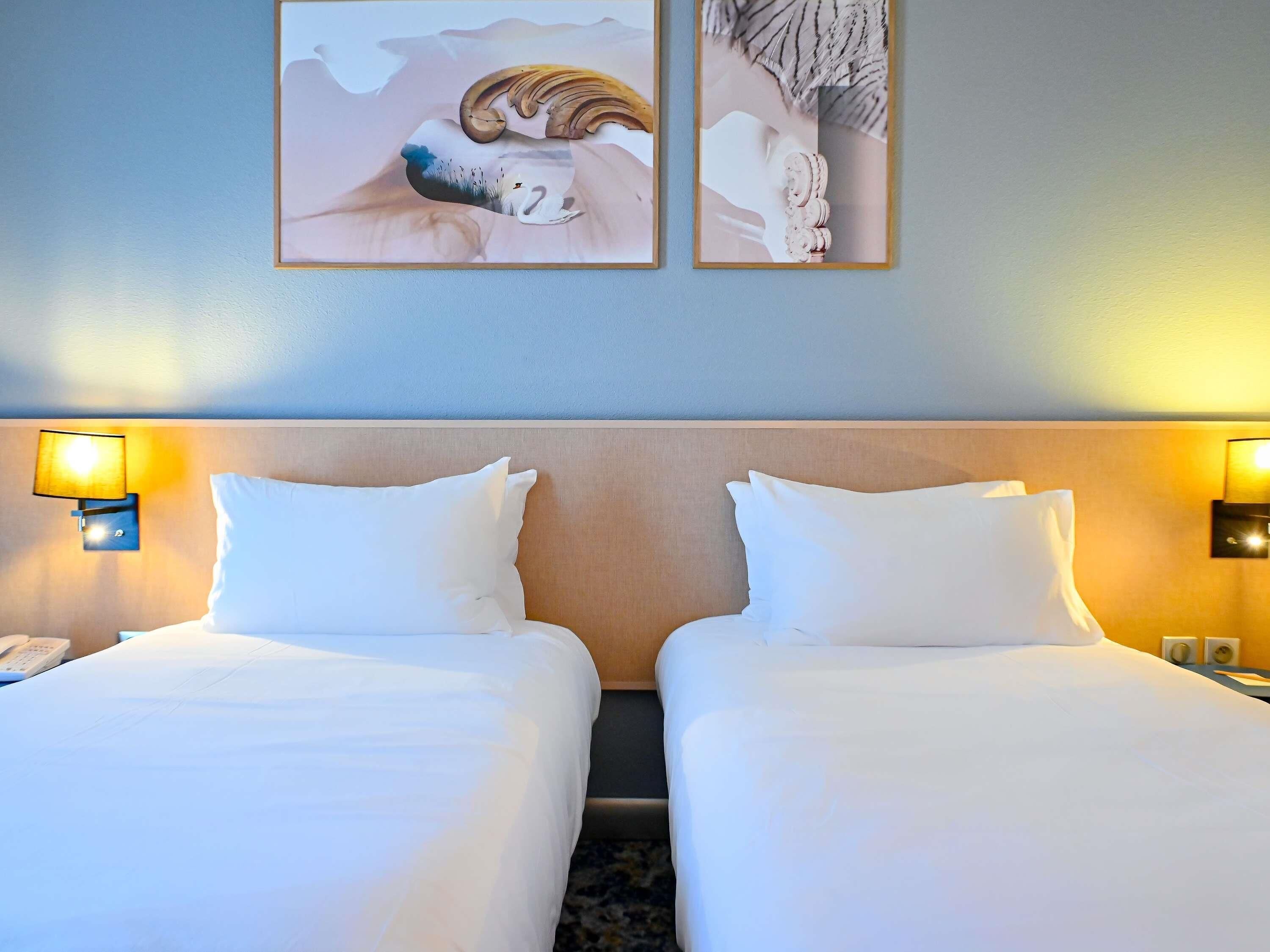 A hotel room with two beds, a wooden headboard, wall-mounted lamps, and two framed artworks.