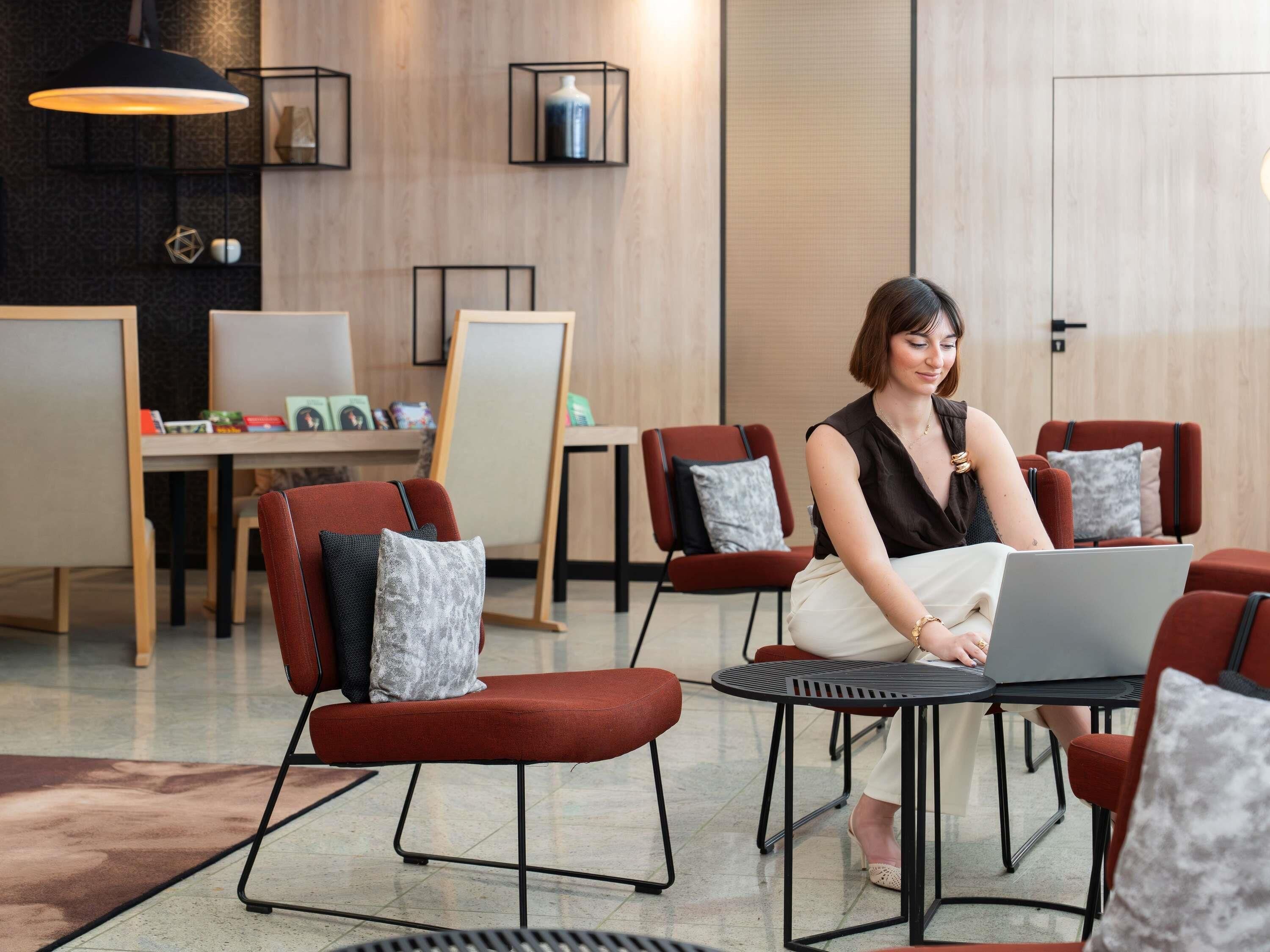 A modern interior with a woman using a laptop at a round table, surrounded by red chairs with grey cushions, in a room with wooden paneling and a large window.