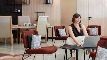A modern interior with a woman using a laptop at a round table, surrounded by red chairs with grey cushions, in a room with wooden paneling and a large window.