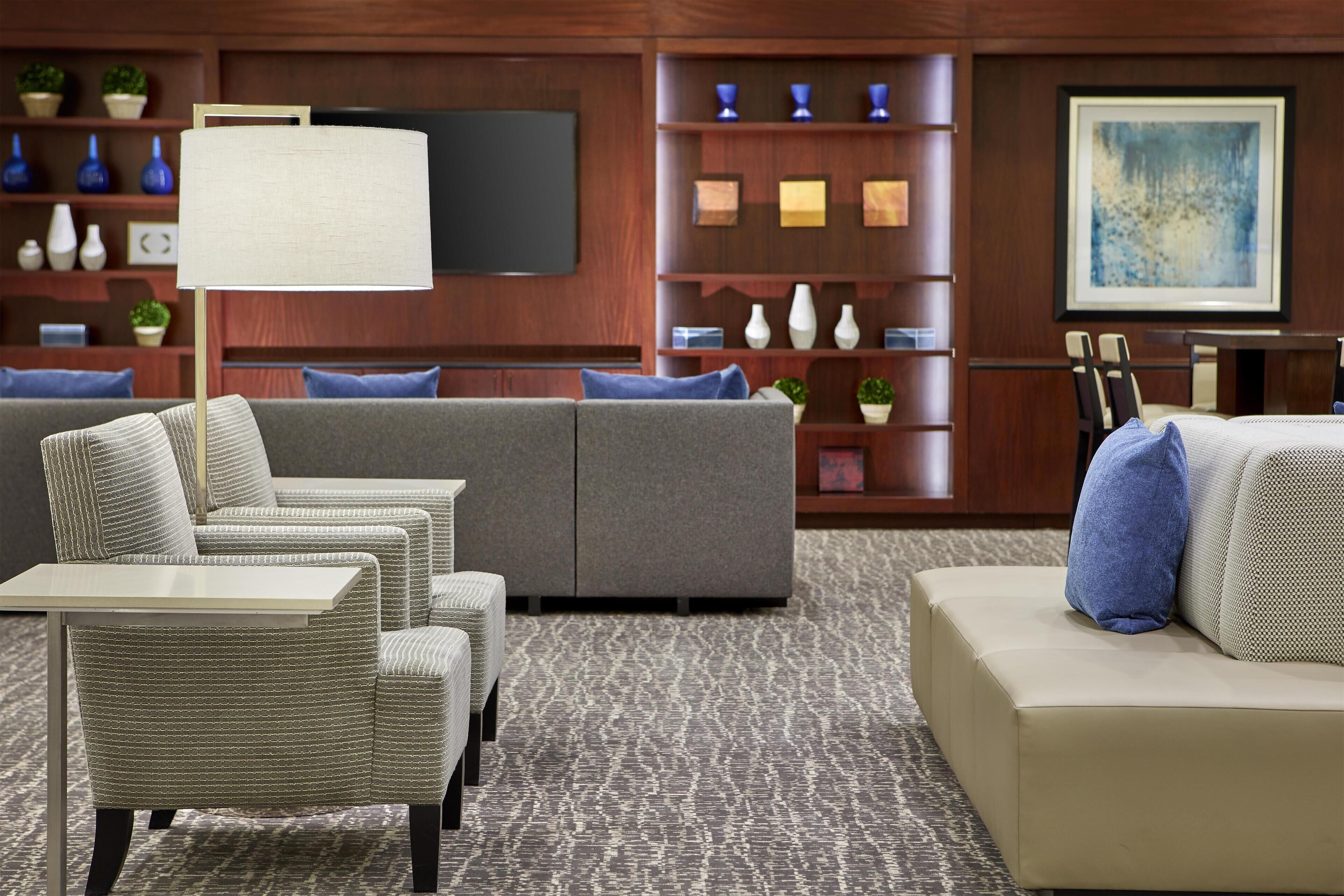 A modern hotel lobby with a grey sofa, wicker chairs, a table, and a large wall-mounted TV.