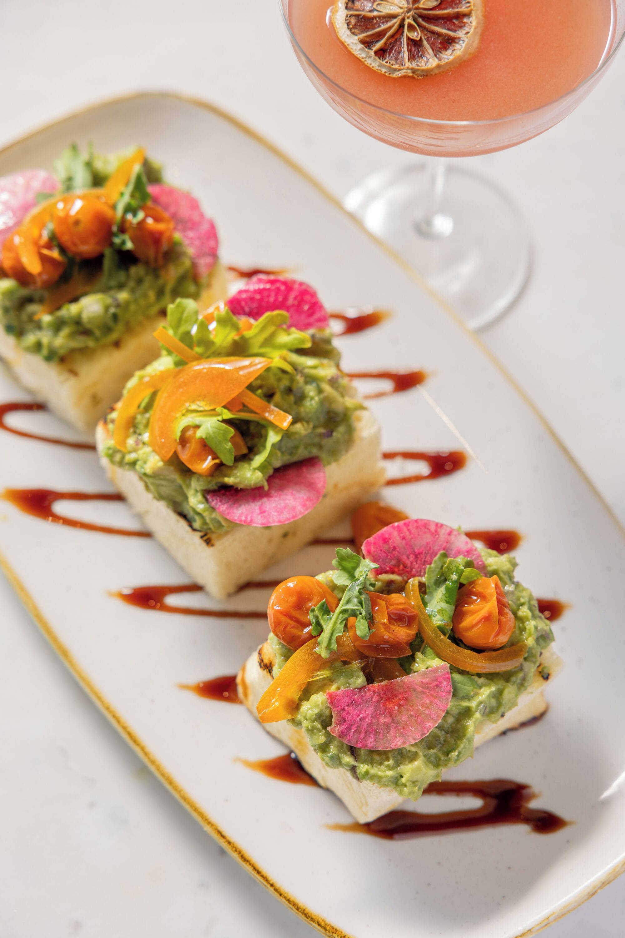 Un piatto con tre tramezzini avocado, guarniti con ravanelli e carote, e un cocktail con una fetta di frutta secca sul bordo.