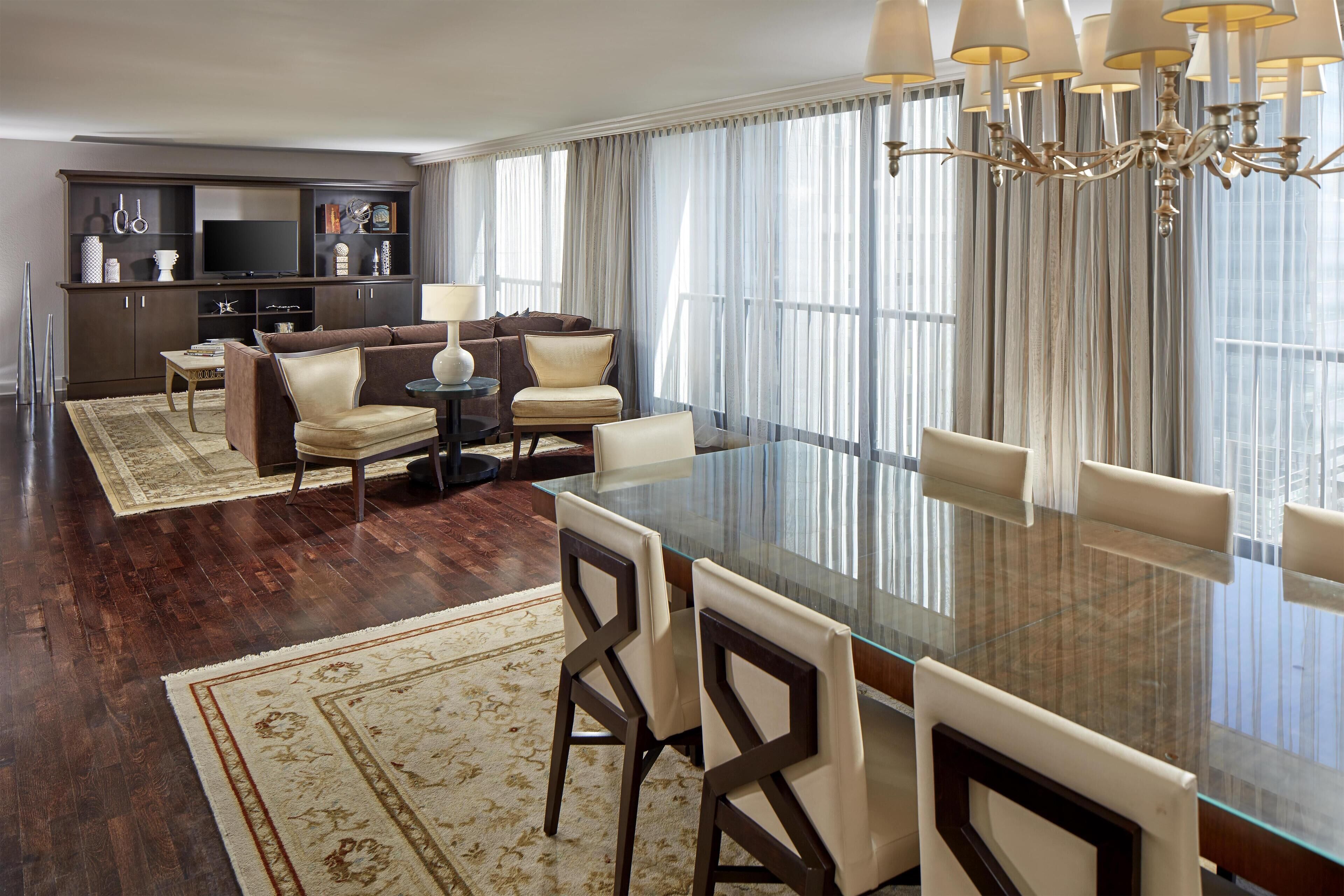 A modern living room with a glass dining table, leather sofas, and a large window with curtains.