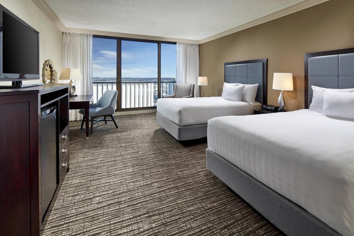 A hotel room with two beds, a desk, a TV, and a view of the city.