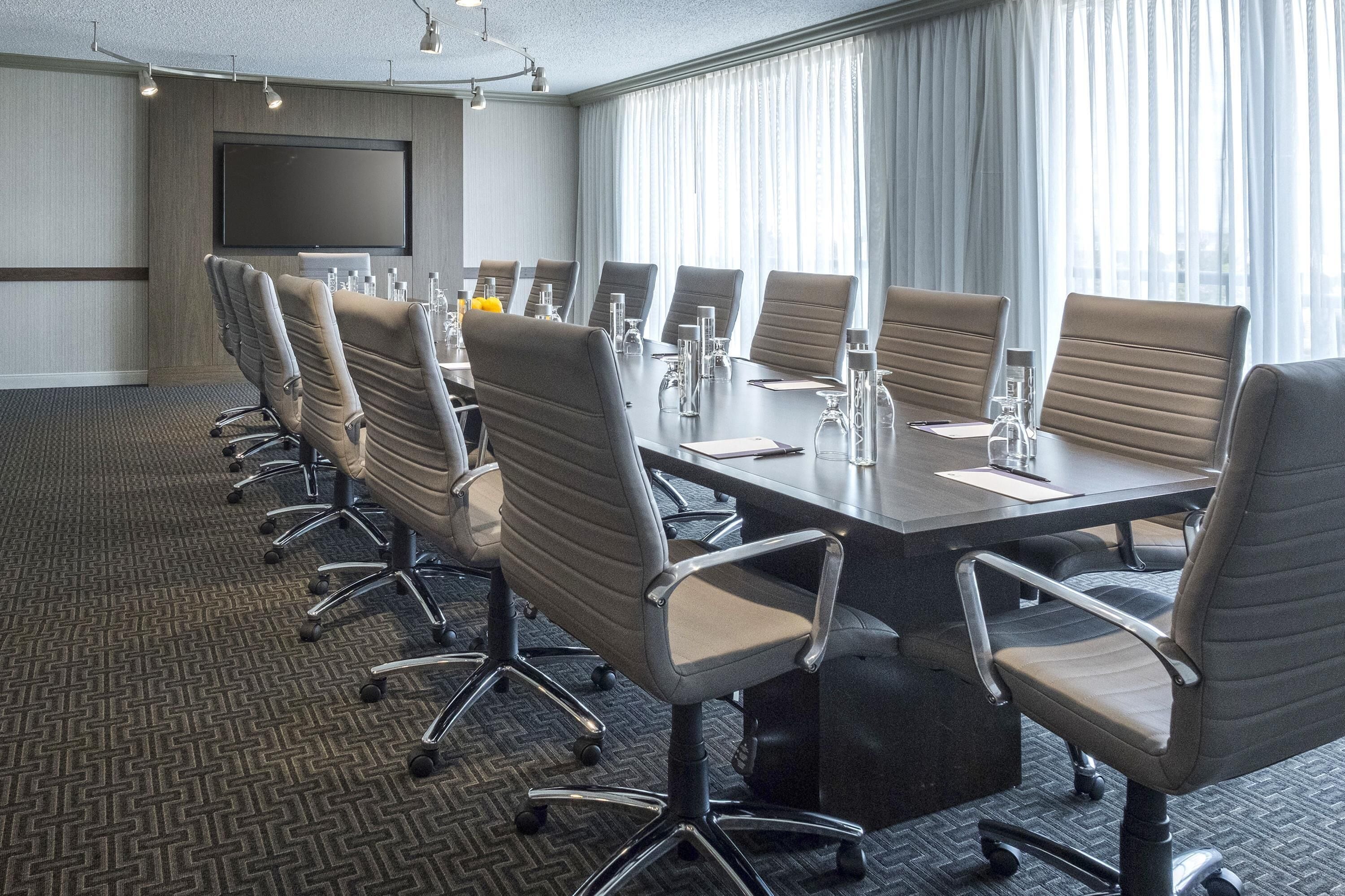A conference room with a large table, multiple chairs, a flat-screen TV, and a large window with curtains.