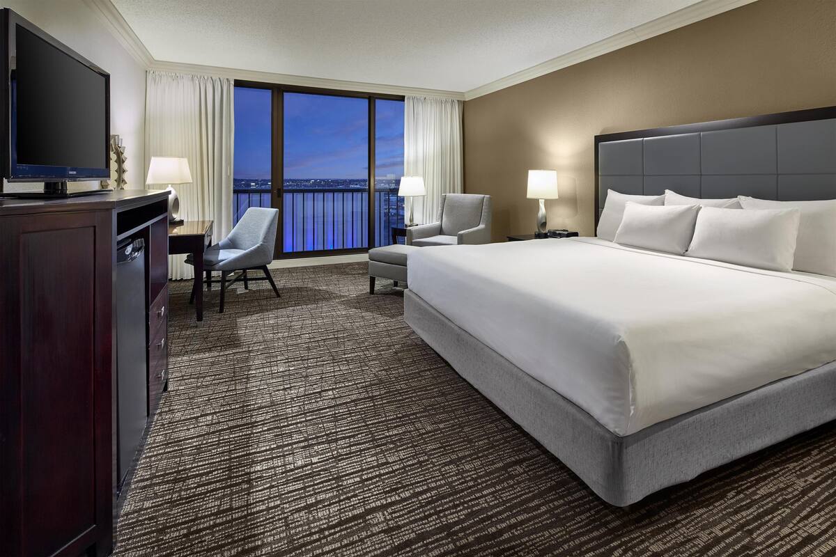 A hotel room with a large bed, a TV, a desk with a chair, and a view of the city at night.