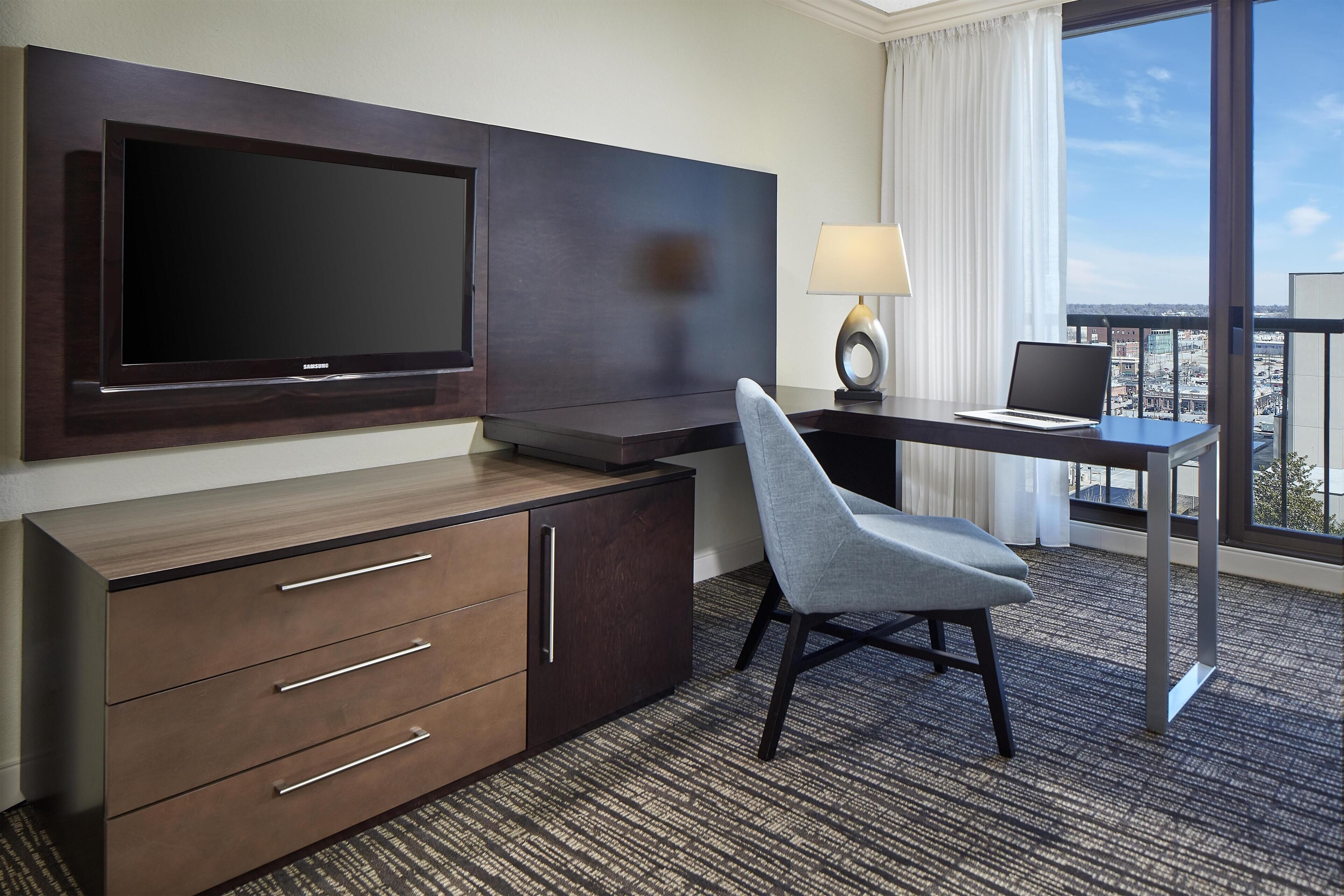 A hotel room with a flat-screen TV mounted on the wall, a desk with a laptop, a chair, a lamp, and a view of the city through large windows.