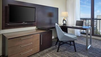A hotel room with a flat-screen TV mounted on the wall, a desk with a laptop, a chair, a lamp, and a view of the city through large windows.