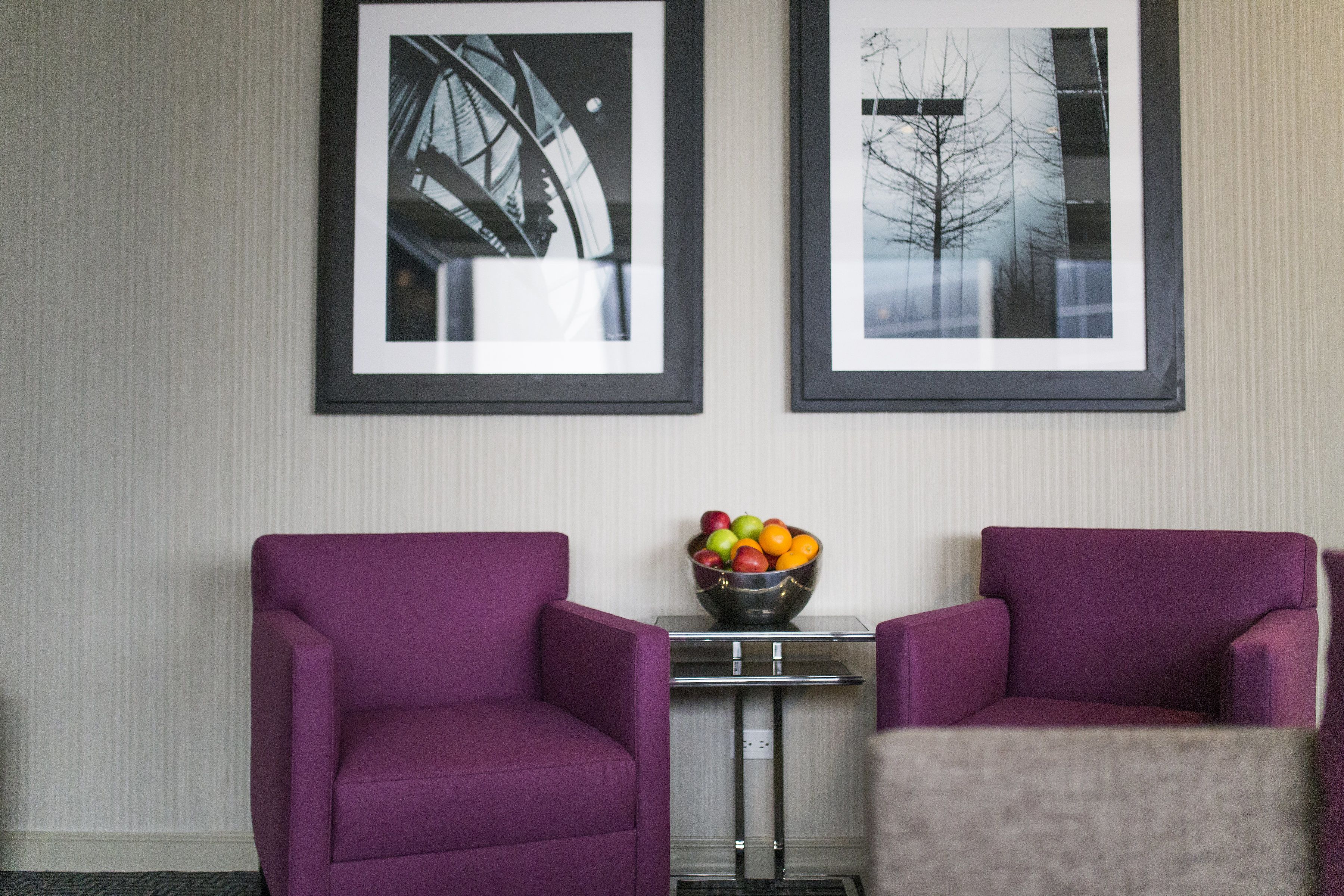 Two purple armchairs, a small table with a fruit bowl, and two framed pictures on the wall.