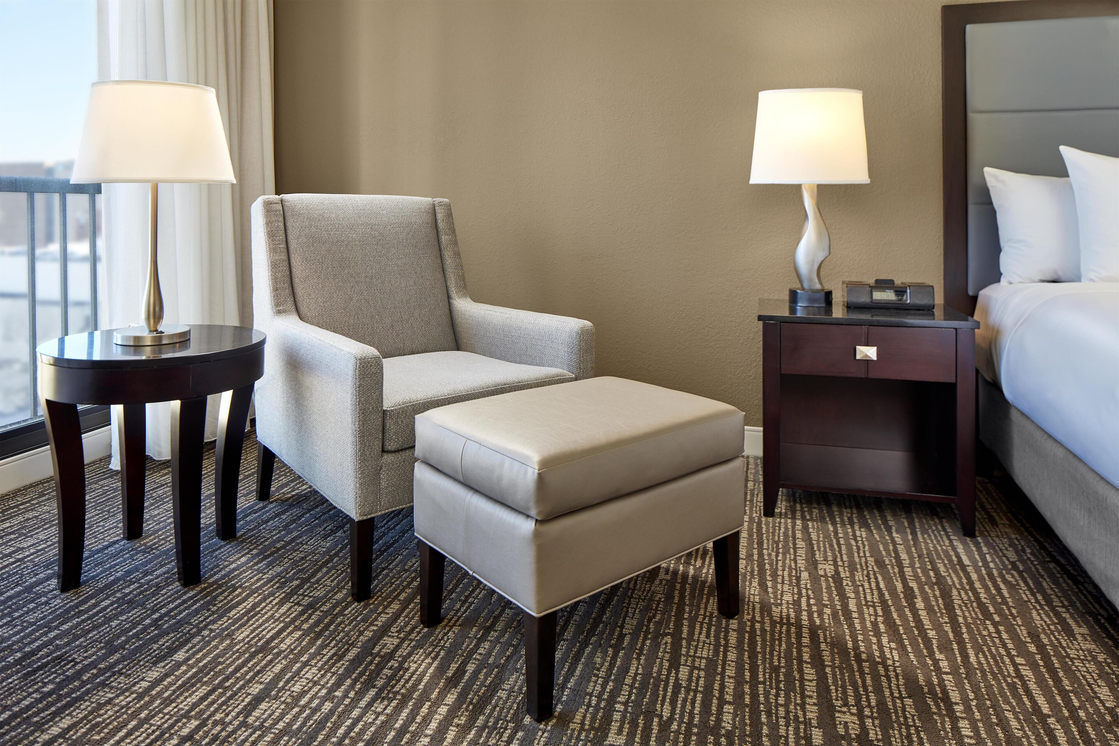 A hotel room with a grey armchair, a matching ottoman, a small table with a lamp, a bedside table with a lamp, and a bed with white bedding.