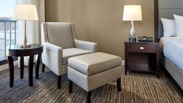 A hotel room with a grey armchair, a matching ottoman, a small table with a lamp, a bedside table with a lamp, and a bed with white bedding.