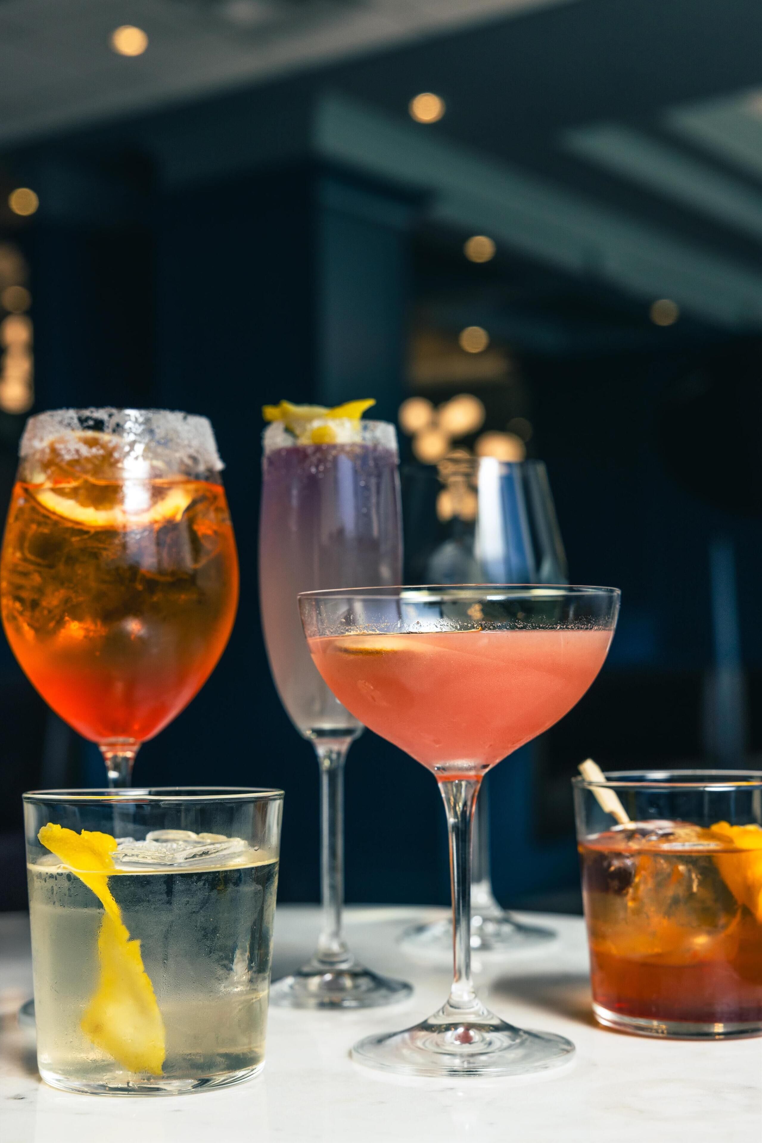 A variety of drinks in different glassware, including a tall glass with ice and a lemon slice, a martini glass, and a short glass with a lemon garnish.
