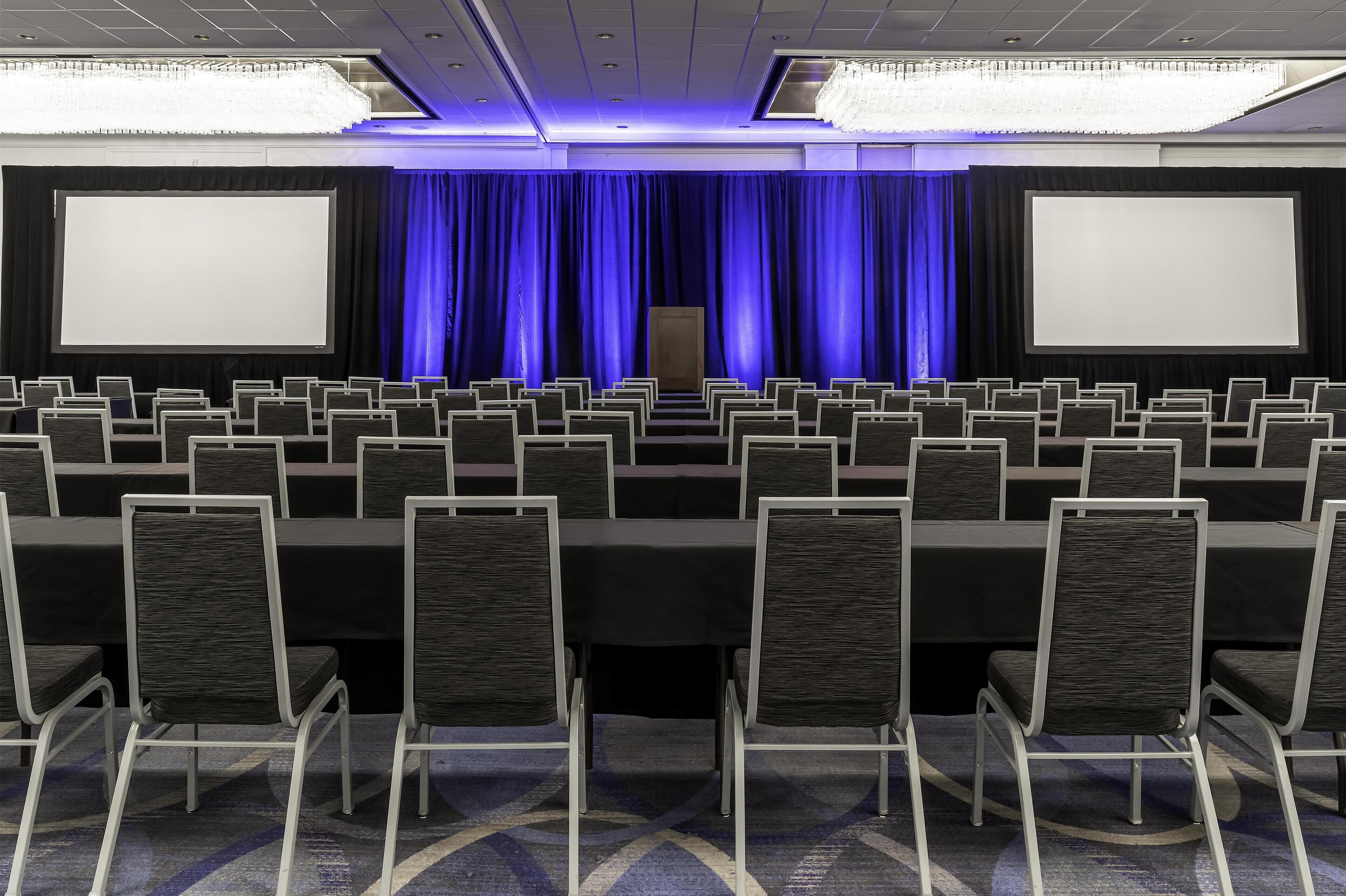 A conference room with rows of chairs, a stage with curtains, and two large screens.
