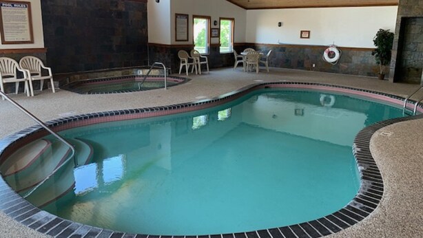 An indoor swimming pool with a tiled edge, a wooden ceiling, and a stone wall. There are chairs and a lifebuoy visible. - Country Inn Deerwood (Deerwood)