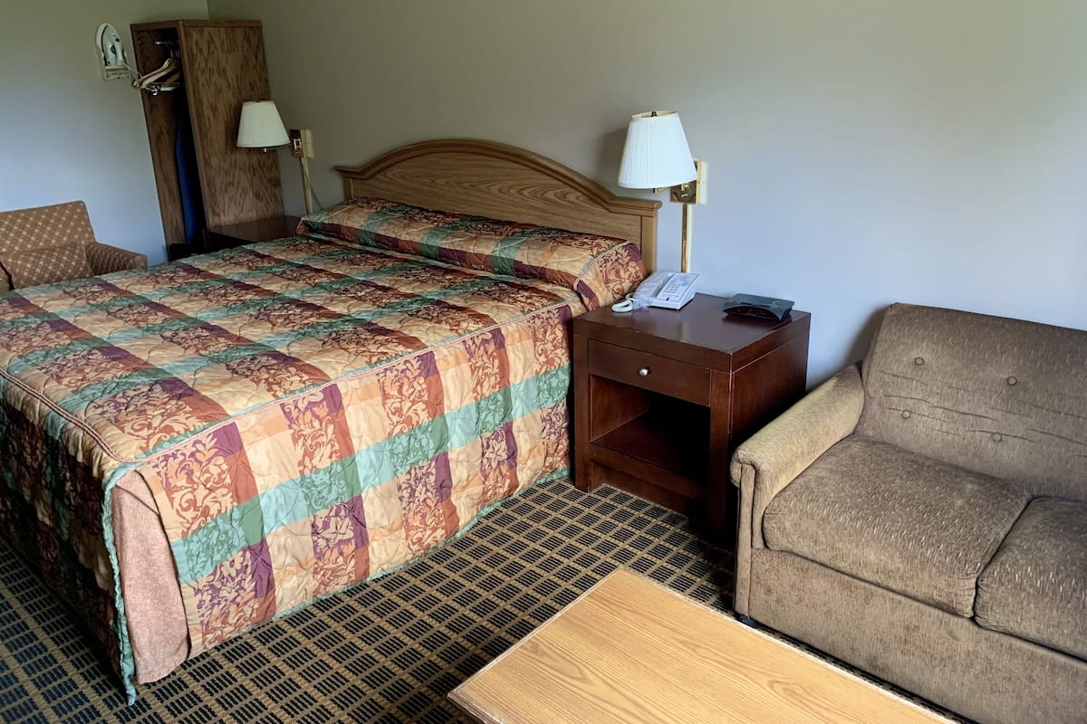 A hotel room with a bed, a wooden nightstand with a lamp, a telephone, and a sofa.