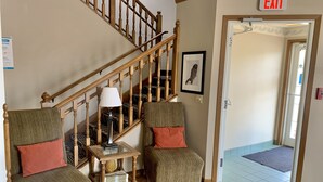 A staircase with wooden railings, two upholstered chairs with cushions, a small table with a lamp, and an exit sign. - Country Inn Deerwood (Deerwood)