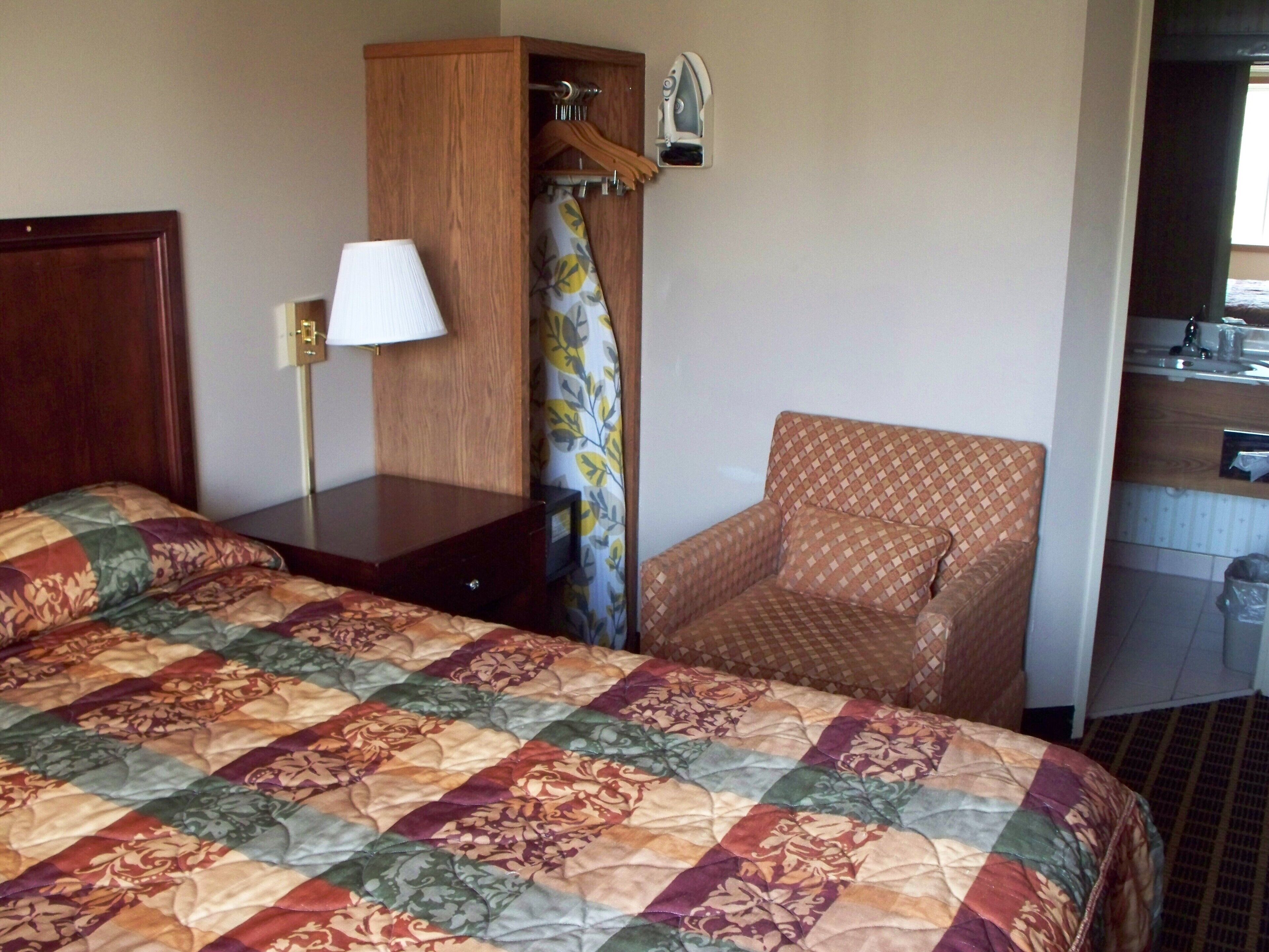 A hotel room with a bed, a chair, a desk with a lamp, and a small kitchenette.