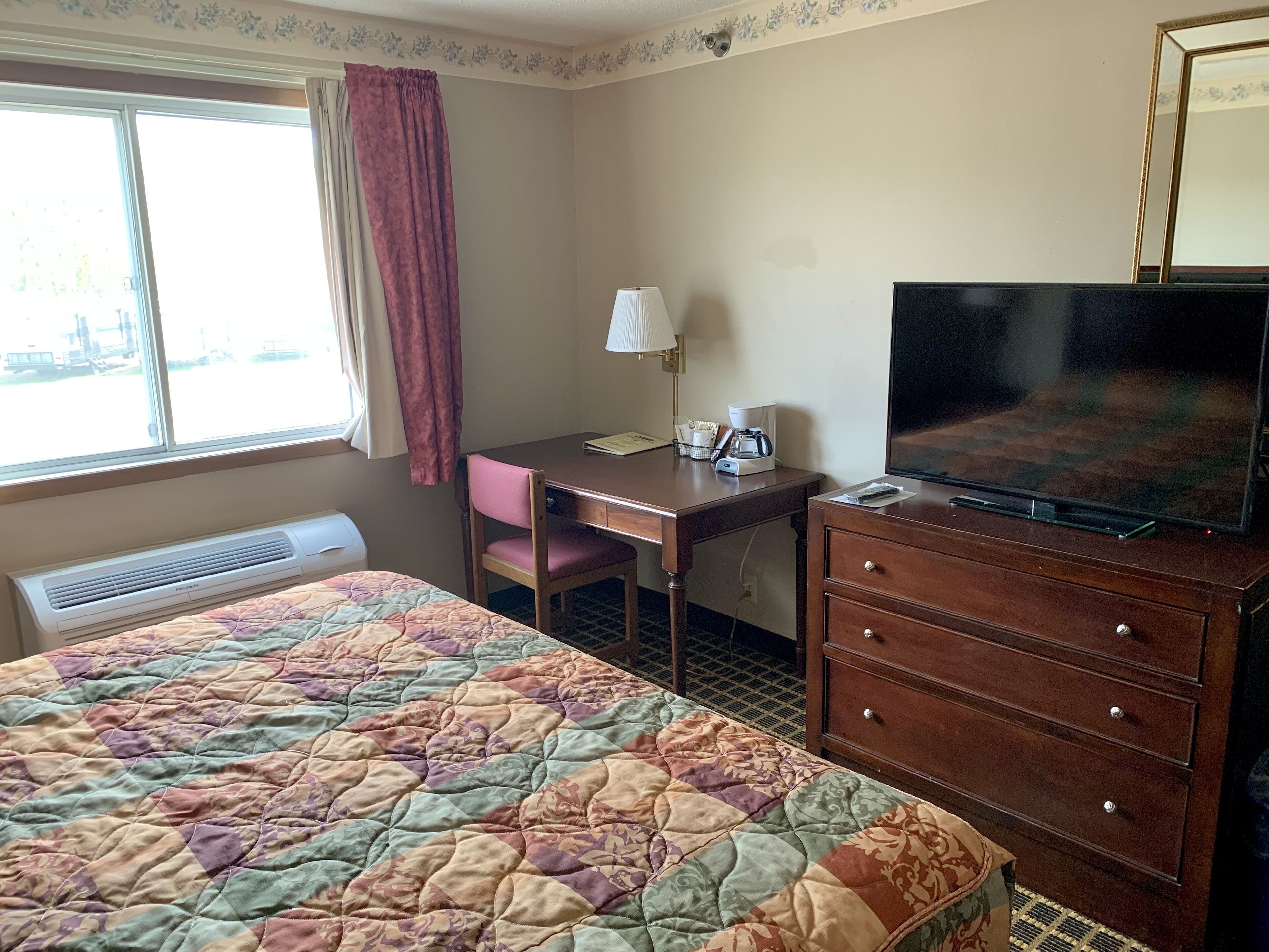 A hotel room with a bed, a desk, a television, a dresser, a window with curtains, and a wall-mounted air conditioner.