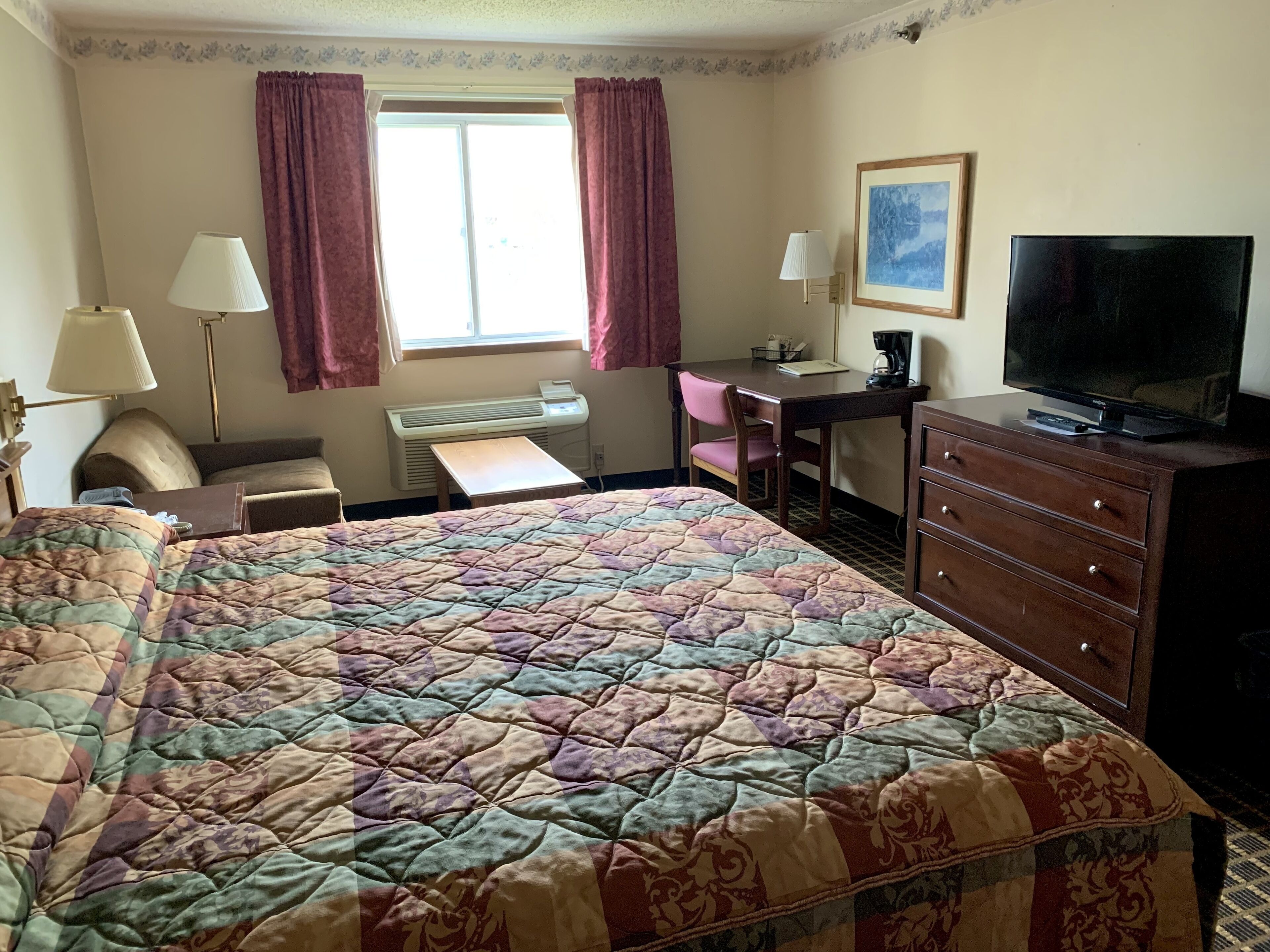 Habitación de hotel con una cama grande, un escritorio, una silla, un aparador, un televisor y una ventana con cortinas.