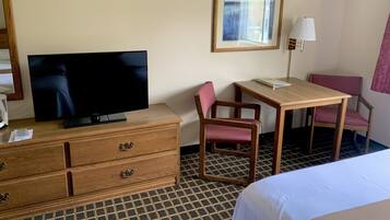 A hotel room with a flat-screen TV on a wooden dresser, a desk with a chair, a framed picture on the wall, and a lamp.