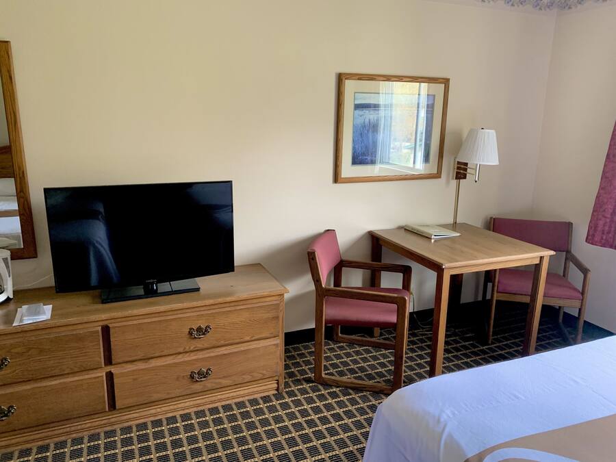 A hotel room with a flat-screen TV on a wooden dresser, a desk with a chair, a framed picture on the wall, and a lamp.
