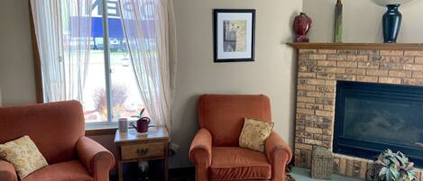 A living room with two armchairs, a small table with a vase, a framed picture, and a brick fireplace.