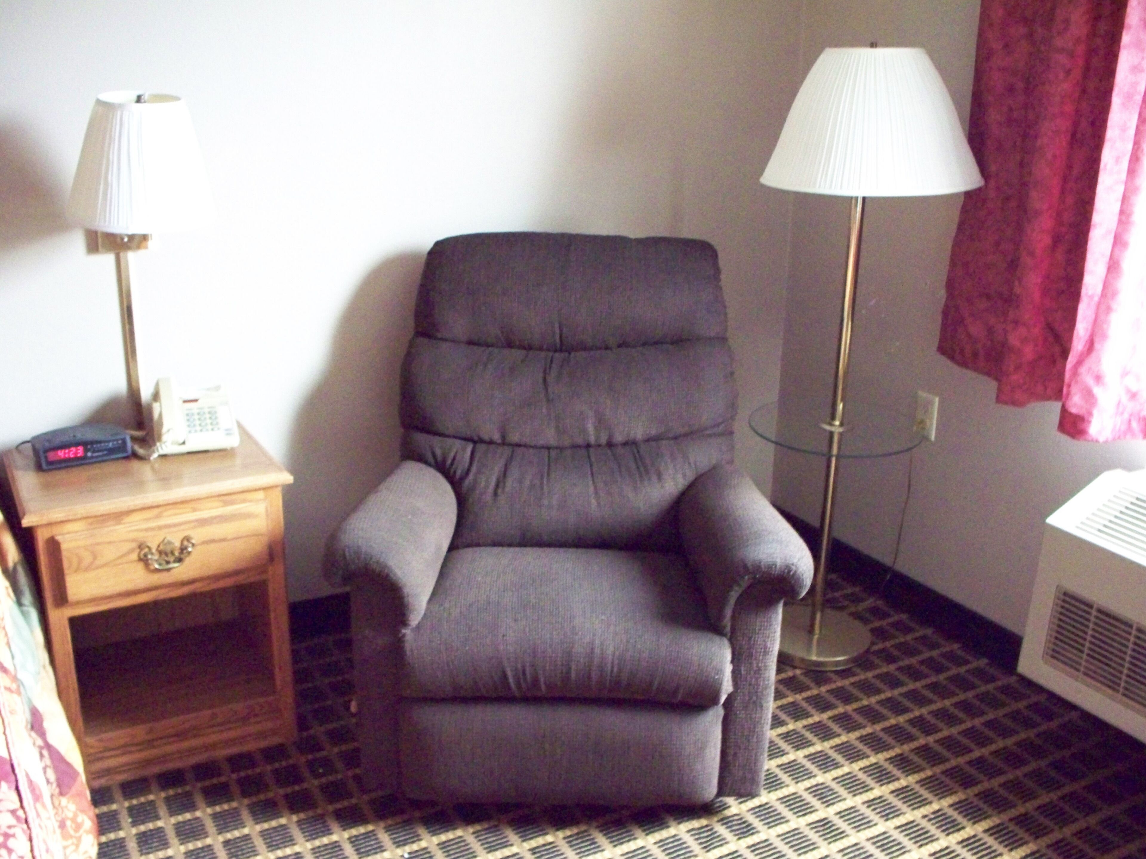 Une pièce avec un fauteuil gris, une table de chevet avec une lampe, un téléphone et un radiateur.