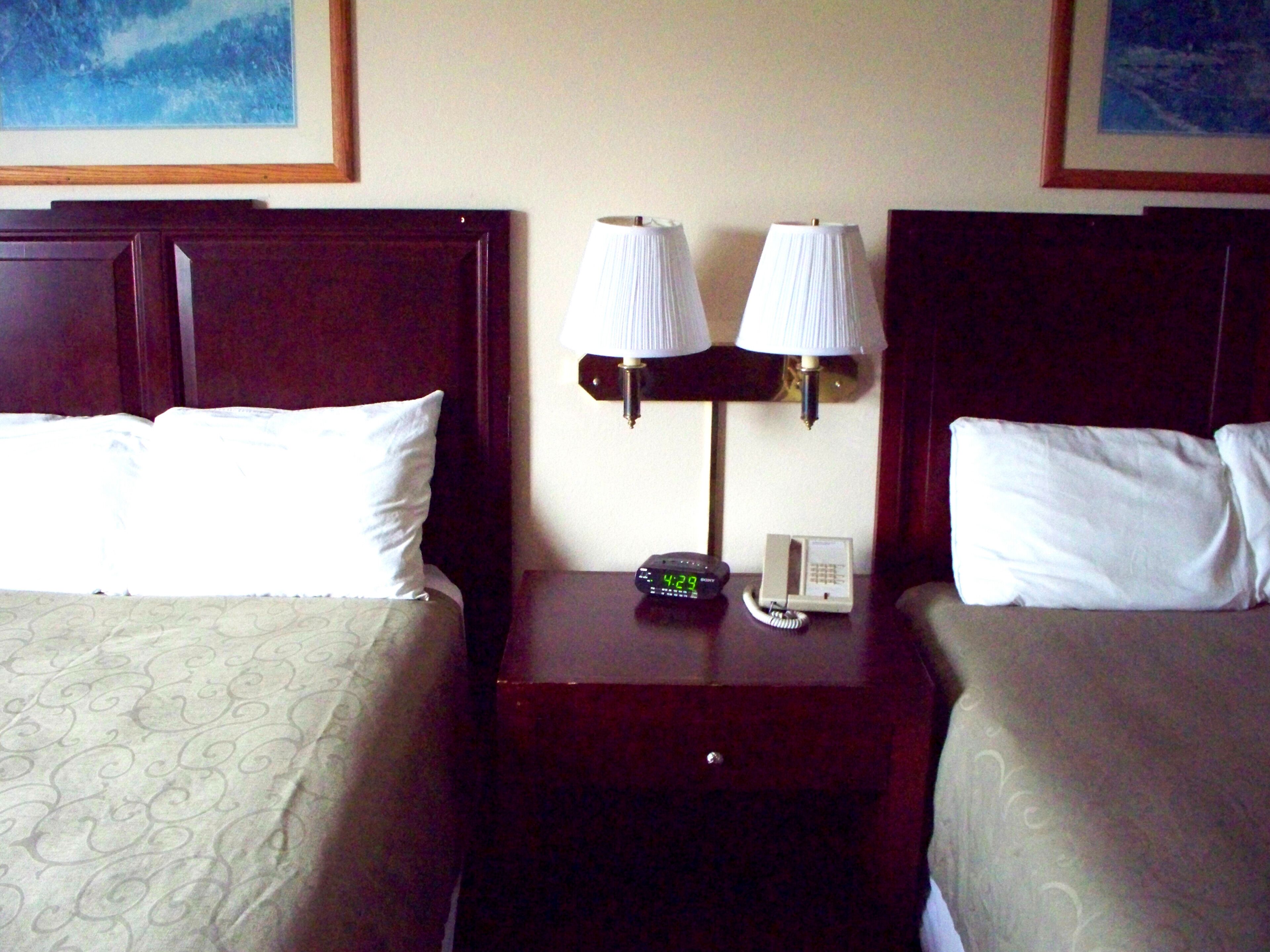 A hotel room with two beds, each with a pillow and a patterned bedspread. There is a nightstand with a lamp, a clock, and a telephone.