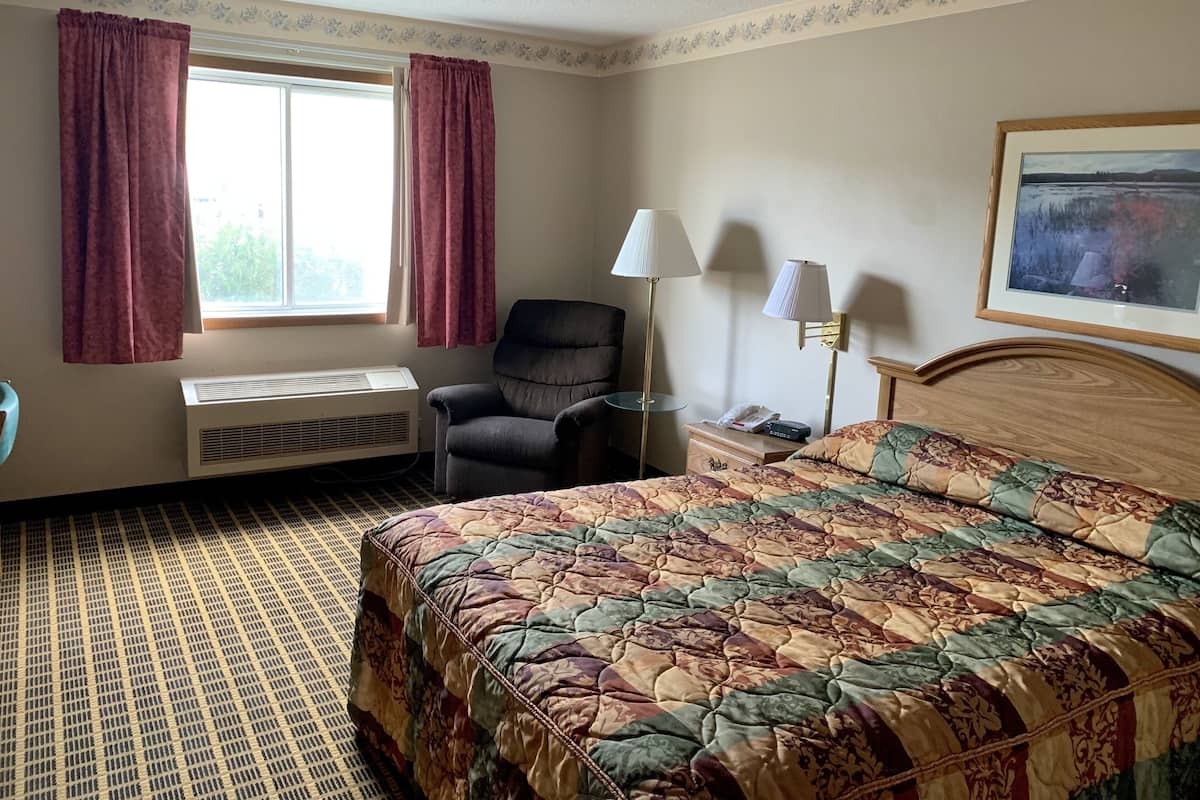 A hotel room with a bed, a chair, a desk with a lamp, a window with curtains, and a radiator.