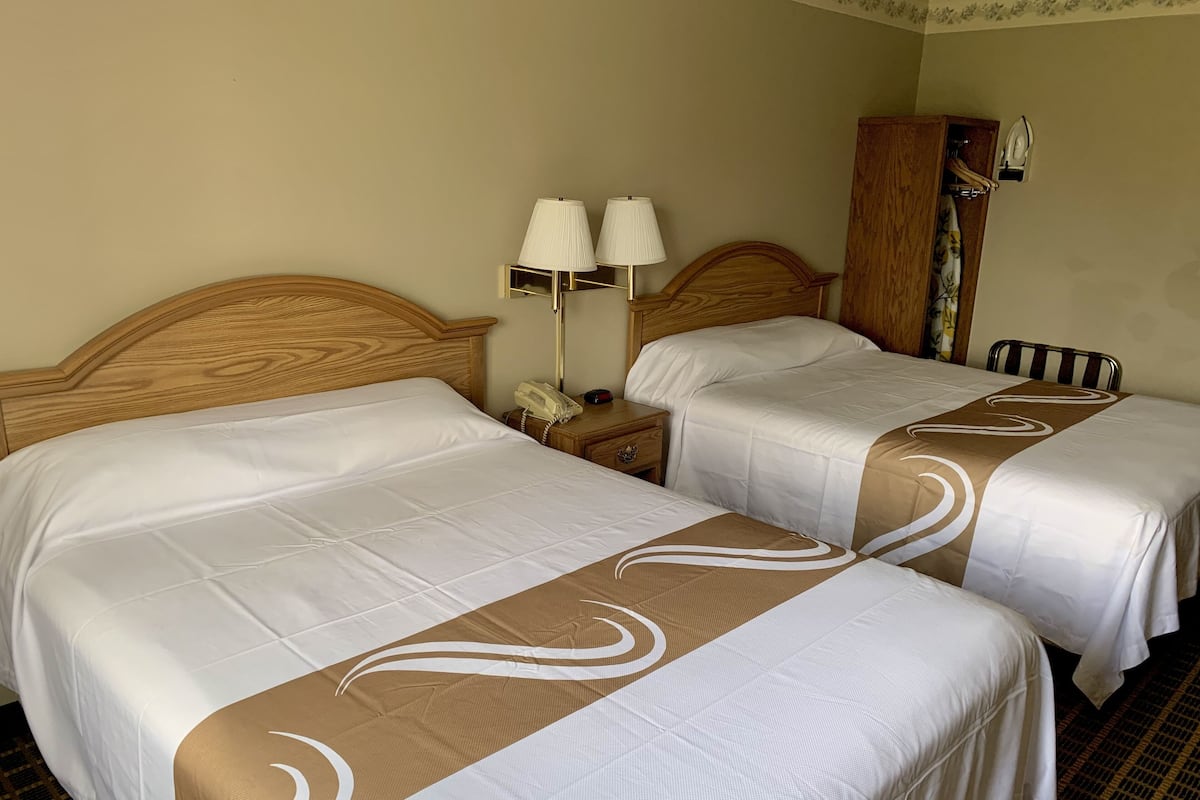 A hotel room with two beds, each with a wooden headboard, white bedding, and a brown decorative runner. There is a nightstand with a lamp and a telephone.
