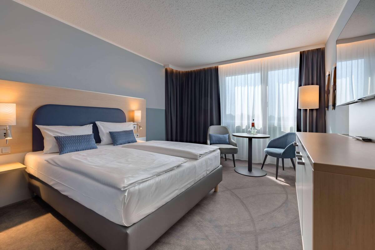 A hotel room with two beds, a desk, a chair, a small table, and a TV.