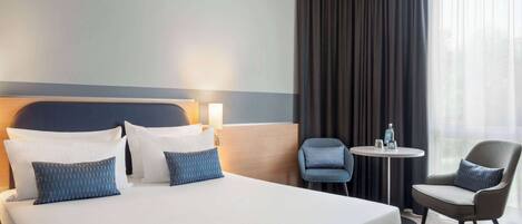 A hotel room with a large bed, two pillows, a bedside table with a lamp, a chair, and a view of the outdoors through a window with curtains.