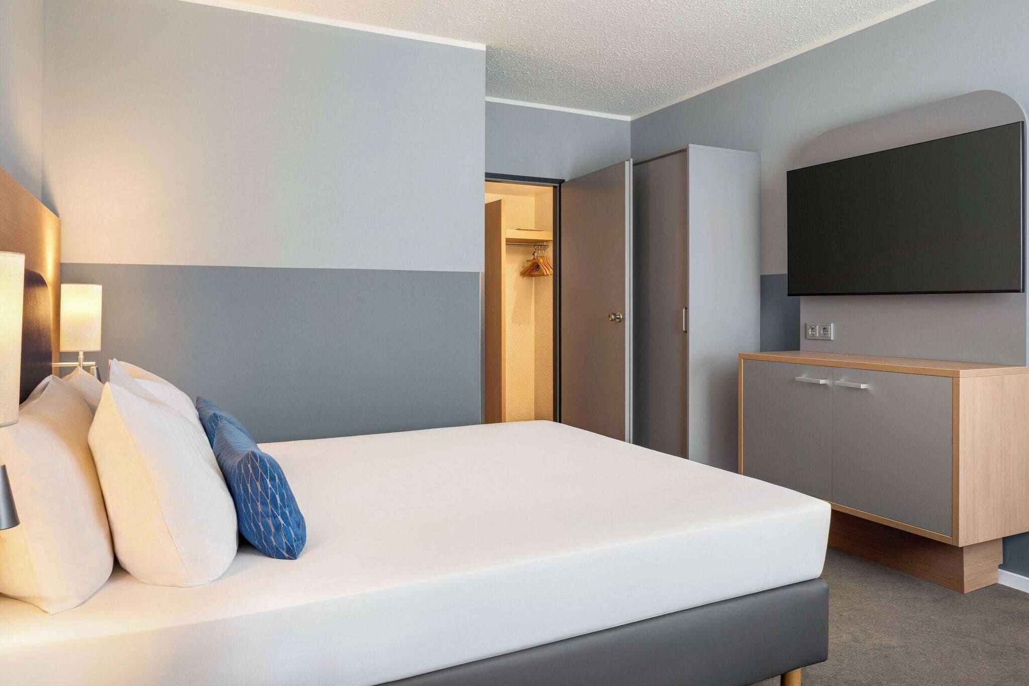 A hotel room with a bed, bedside lamps, a wardrobe, and a flat-screen TV on the wall.