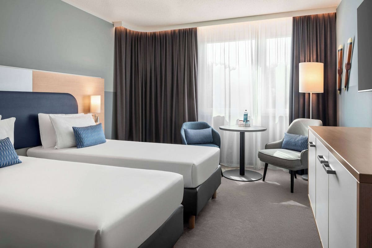 A hotel room with two beds, a desk, a chair, a small table, and a lamp.