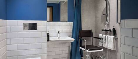A bathroom with a white toilet, a sink, a shower with a handheld showerhead, and a wheelchair.