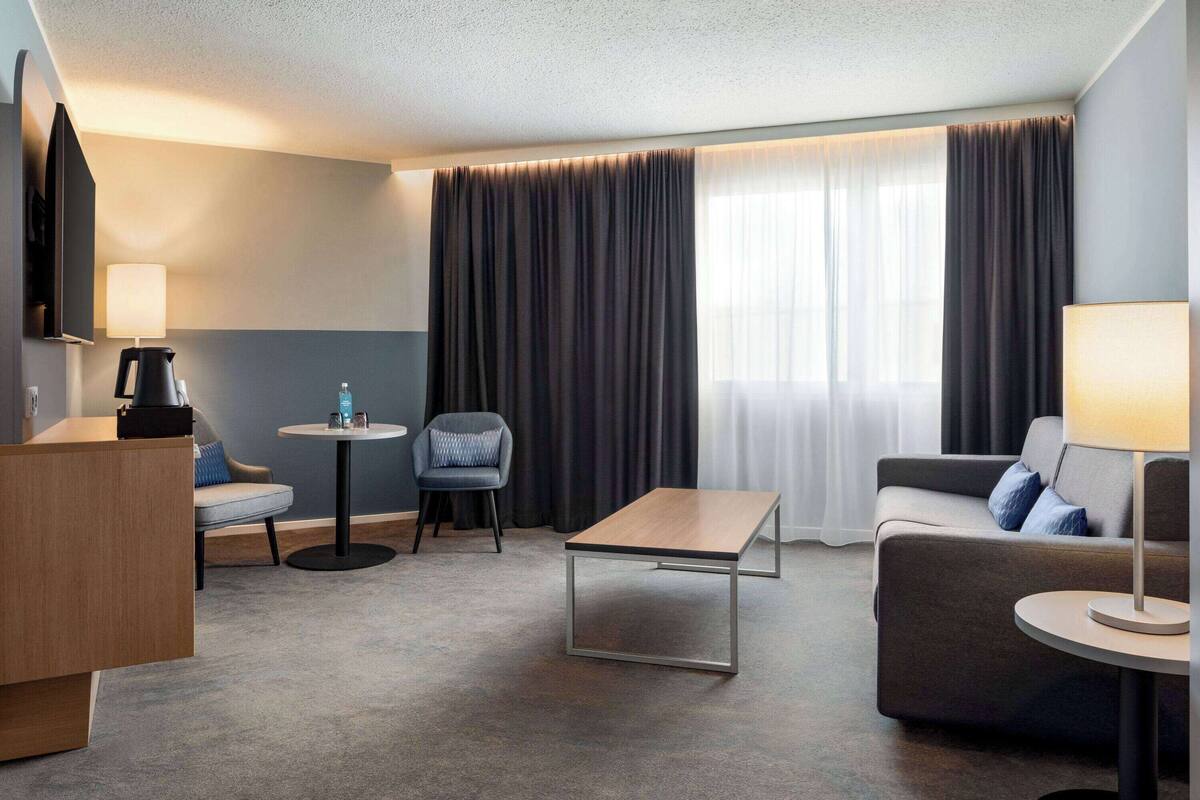 A modern hotel room with a flat-screen TV, a wooden desk, a round coffee table, a sofa, and a chair.