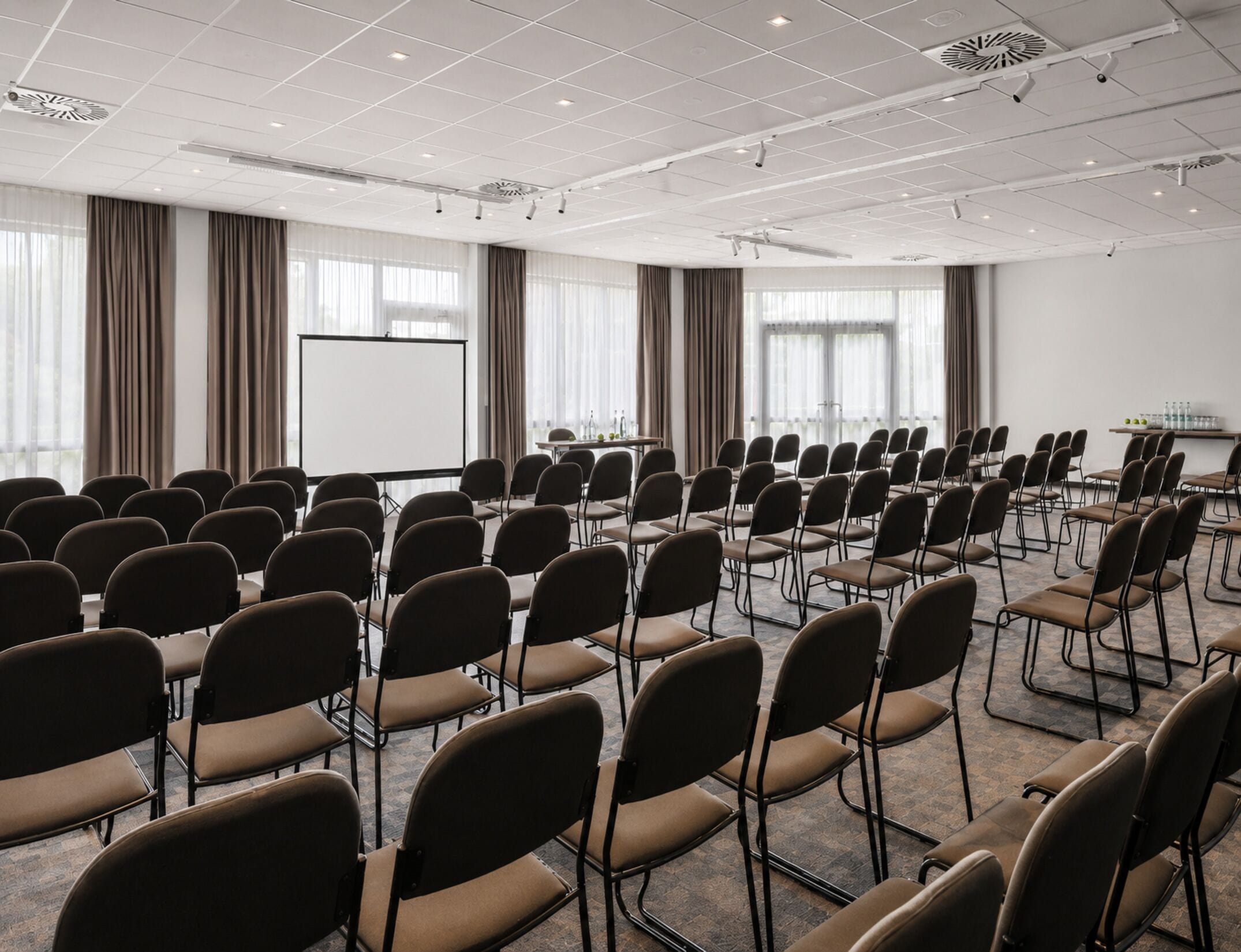 Ein Konferenzraum mit Stuhlreihen, einer Projektionsleinwand und großen Fenstern mit Vorhängen.