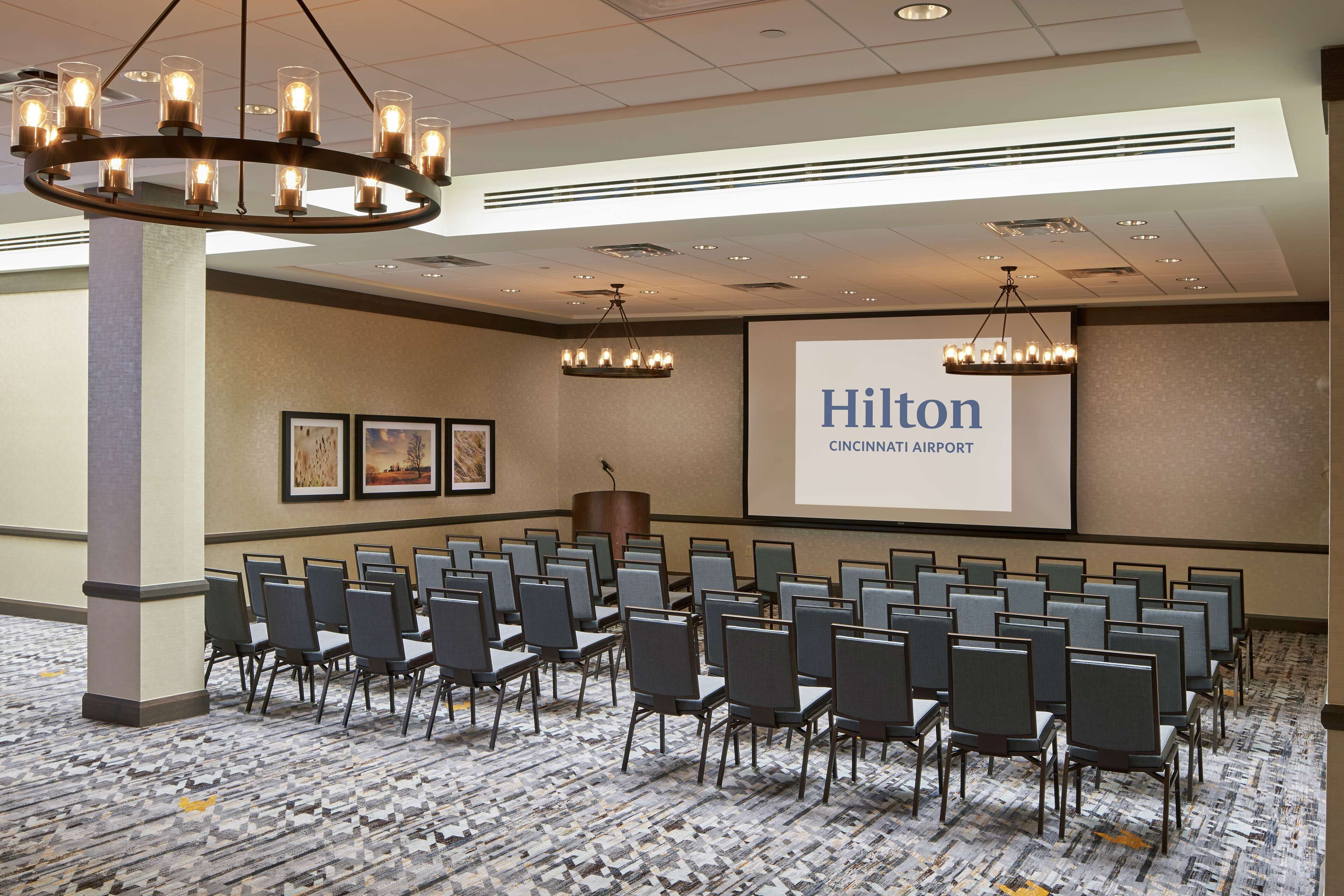 Ett konferensrum med rader av stolar, en projektorduk som visar texten ”Hilton Cincinnati Airport” och en kristallkrona.