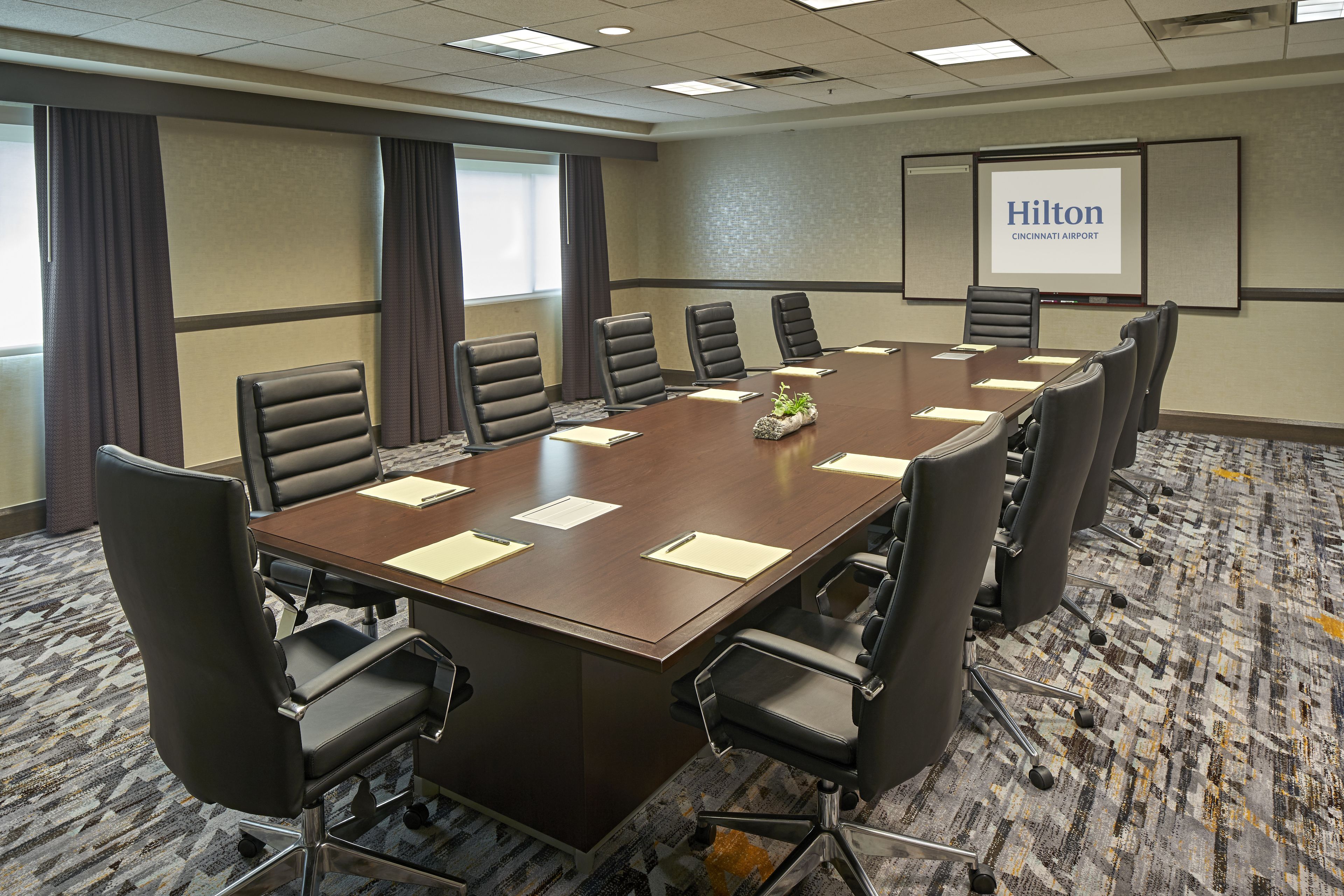 A conference room with a large table, multiple chairs, a projector screen displaying 'Hilton Cincinnati Airport', and a carpeted floor.