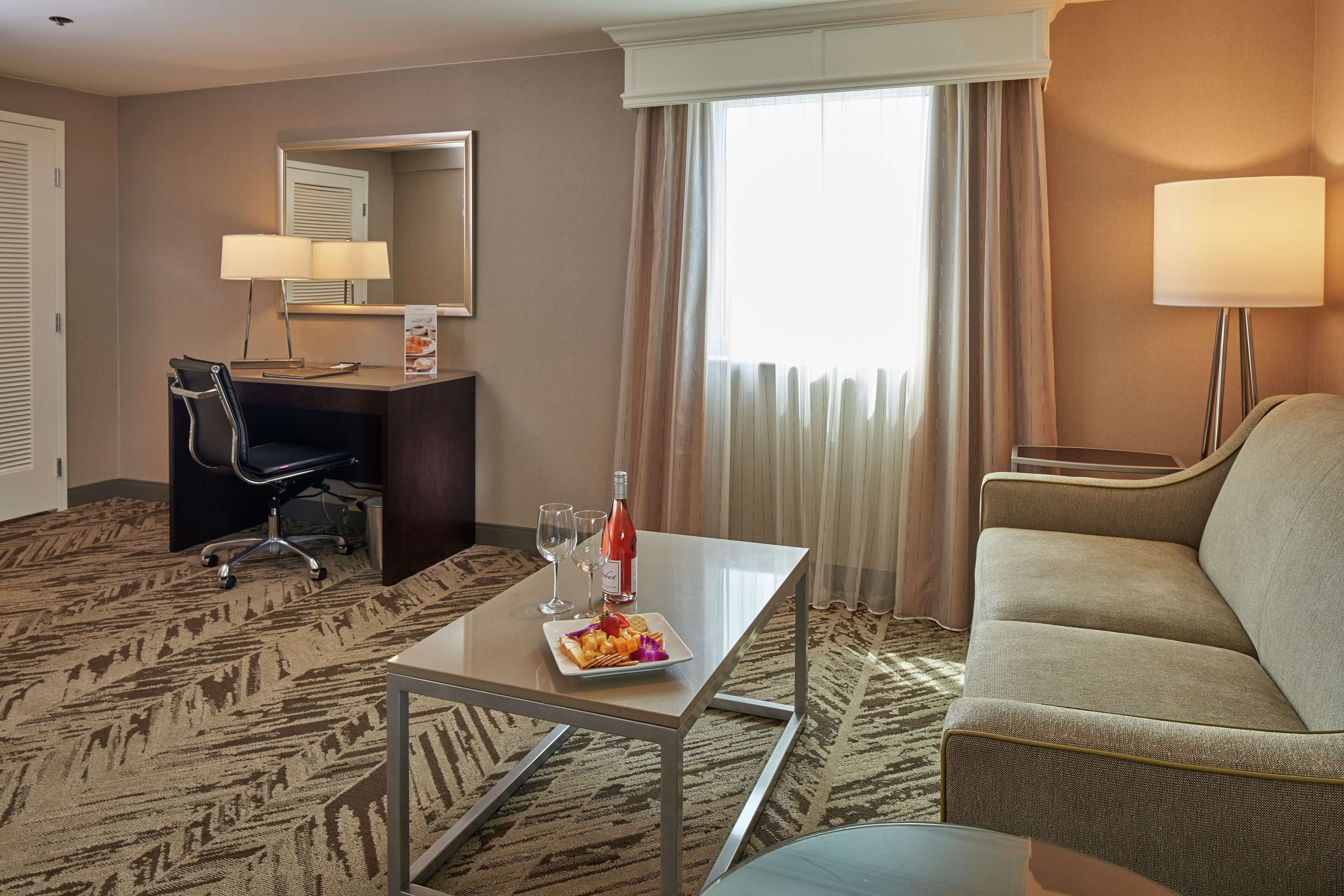 A hotel room with a desk, a chair, a sofa, a table with a plate of fruit and a bottle of wine, and a large window with curtains.