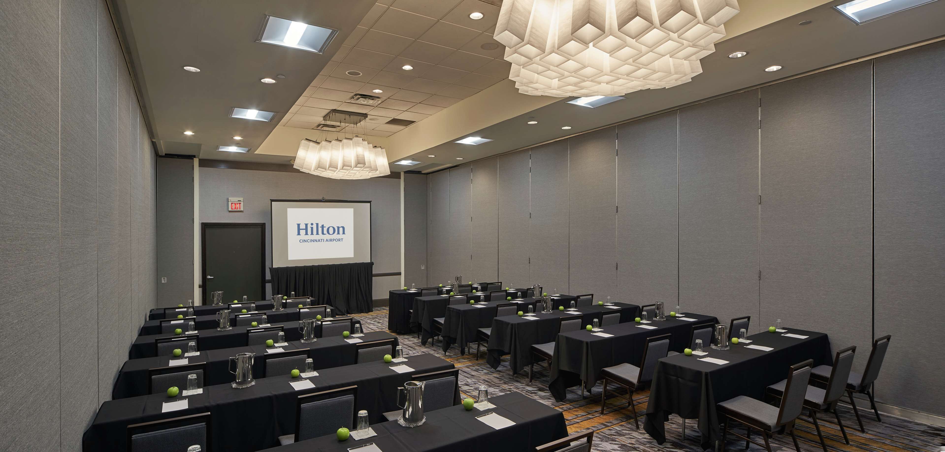 A conference room with multiple tables, chairs, and a projector screen displaying "Hilton Cincinnati Airport".